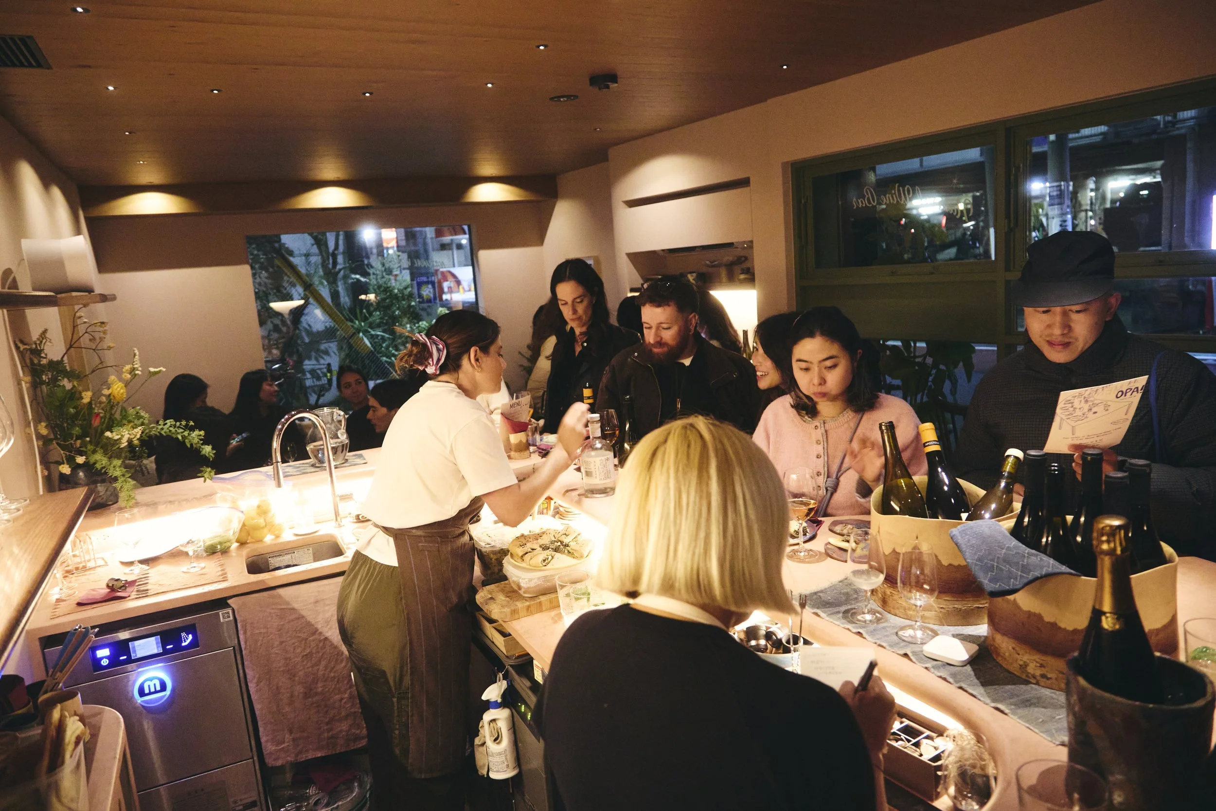 A group of people gathered inside a restaurant or bar, with some standing and others sitting, engaging in conversation and enjoying drinks. There are bottles of wine and glasses on the counter, and a chef or bartender preparing food or drinks. The at