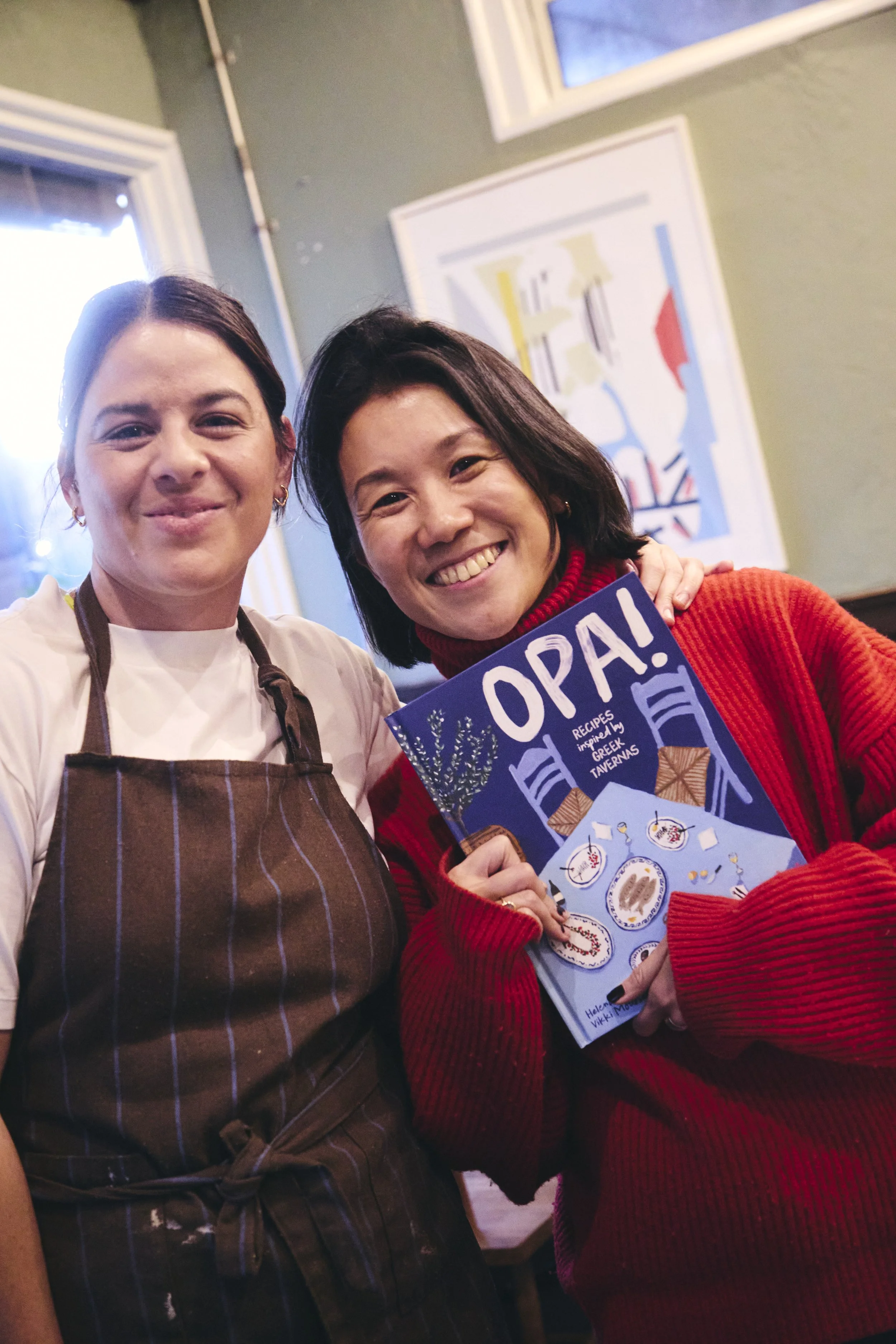 Two smiling women taking a selfie inside a restaurant. One woman is wearing a brown striped apron, and the other woman is holding a menu titled "OPAI" with illustrations of traditional Greek dishes. The background features a window and a colorful fra