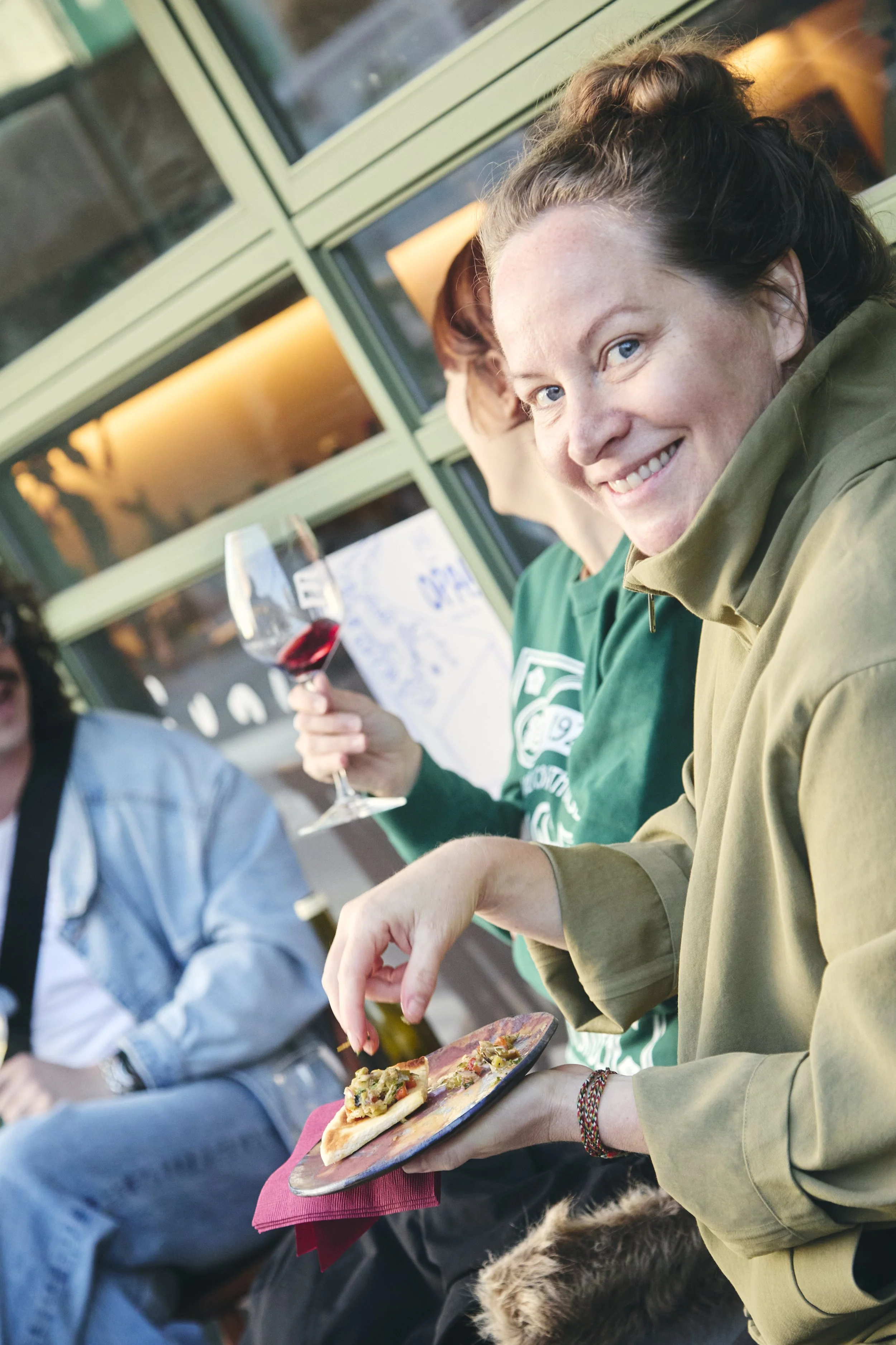 A young woman with curly hair and blue eyes smiling while holding a plate of food at an outdoor gathering. She has a bracelet on her wrist and is wearing a light-colored jacket. Behind her, a person is holding a glass of red wine. The setting appears