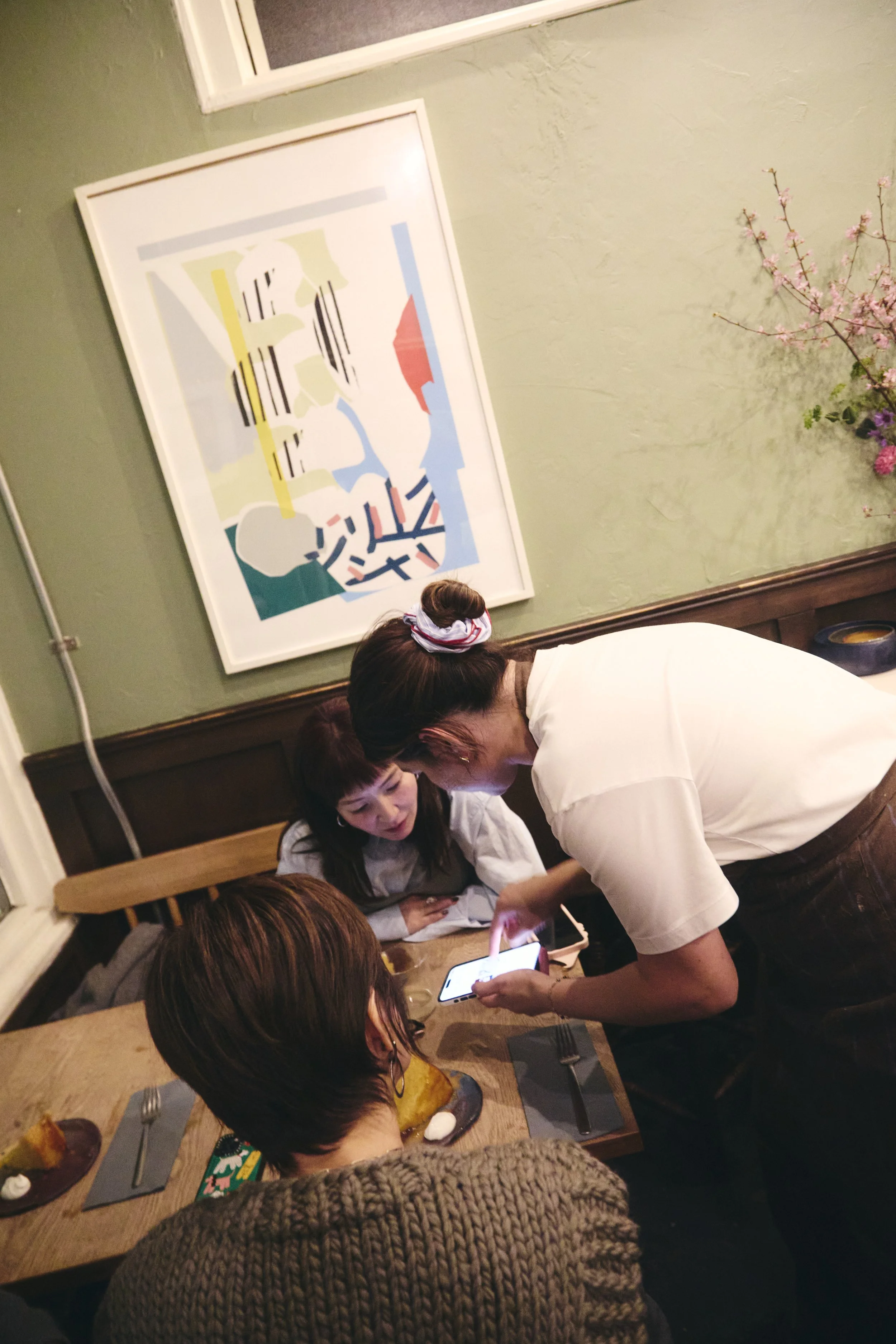 People sitting at a restaurant table with various desserts, a person in a white shirt leaning over, a woman with dark hair looking at a phone, and another person with glasses and a mustard sweater in the foreground. A colorful abstract painting hangs