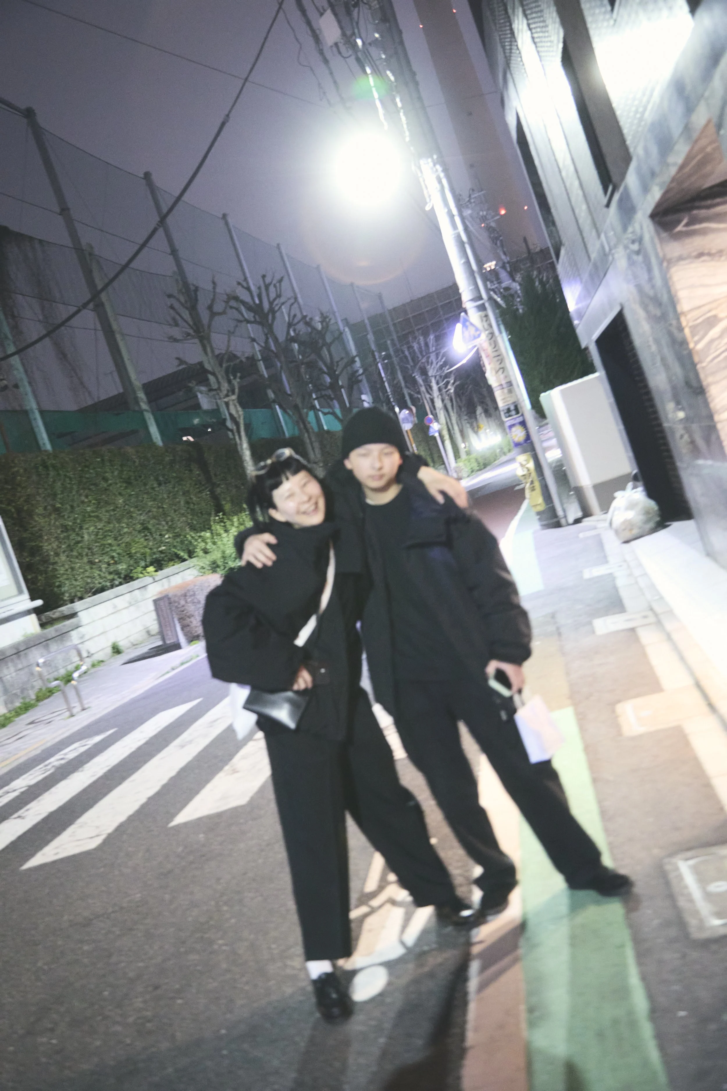 Two people, a woman and a man, are smiling and embracing each other on a street at night. They are dressed warmly in black jackets, with the woman wearing sunglasses on her head and the man wearing a beanie. The street has white pedestrian crossing l
