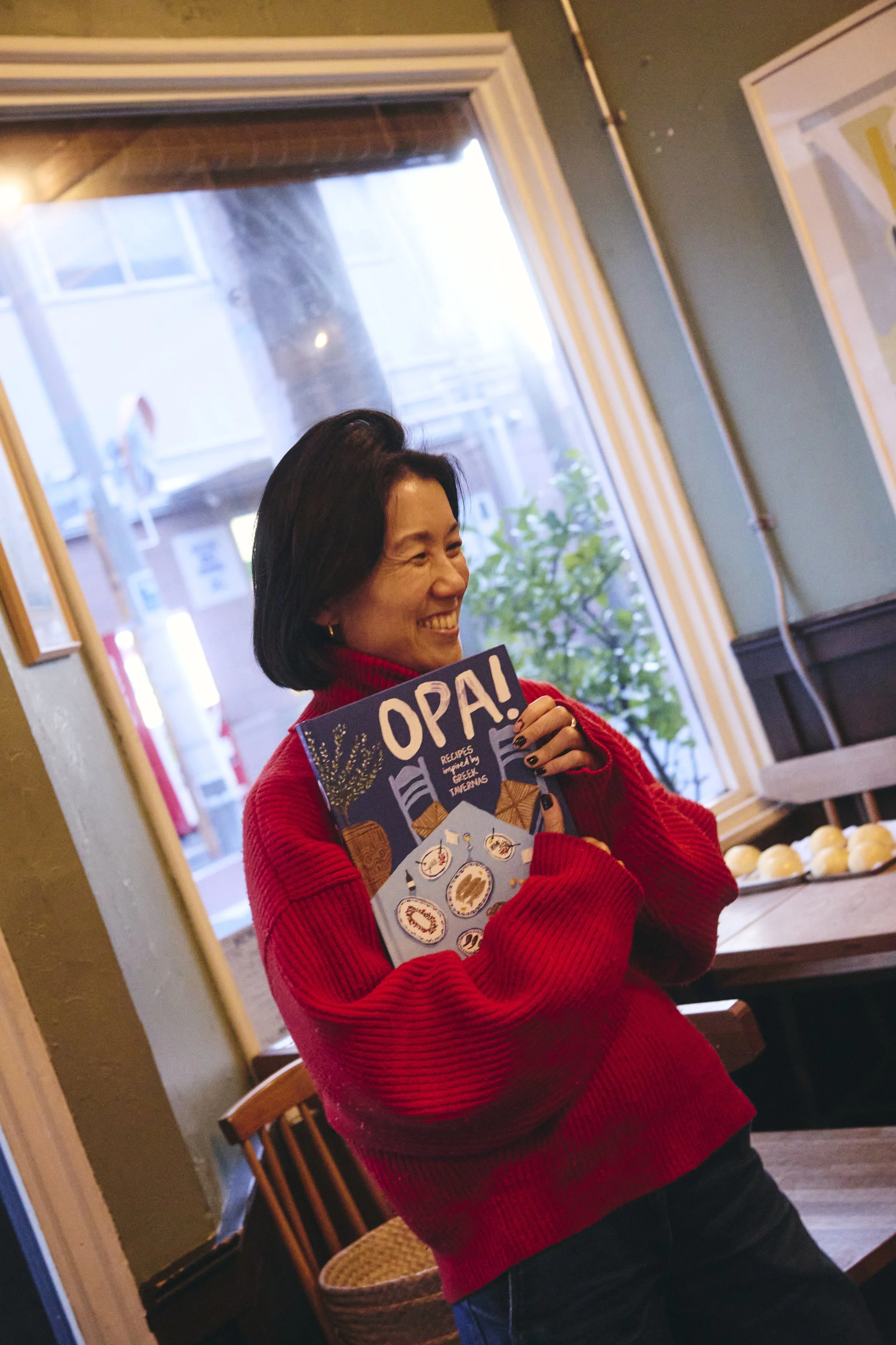 A woman with short dark hair in a red sweater smiling and holding a book titled 'OPAL!' in a cozy indoor setting, with a window and a wooden chair in the background.