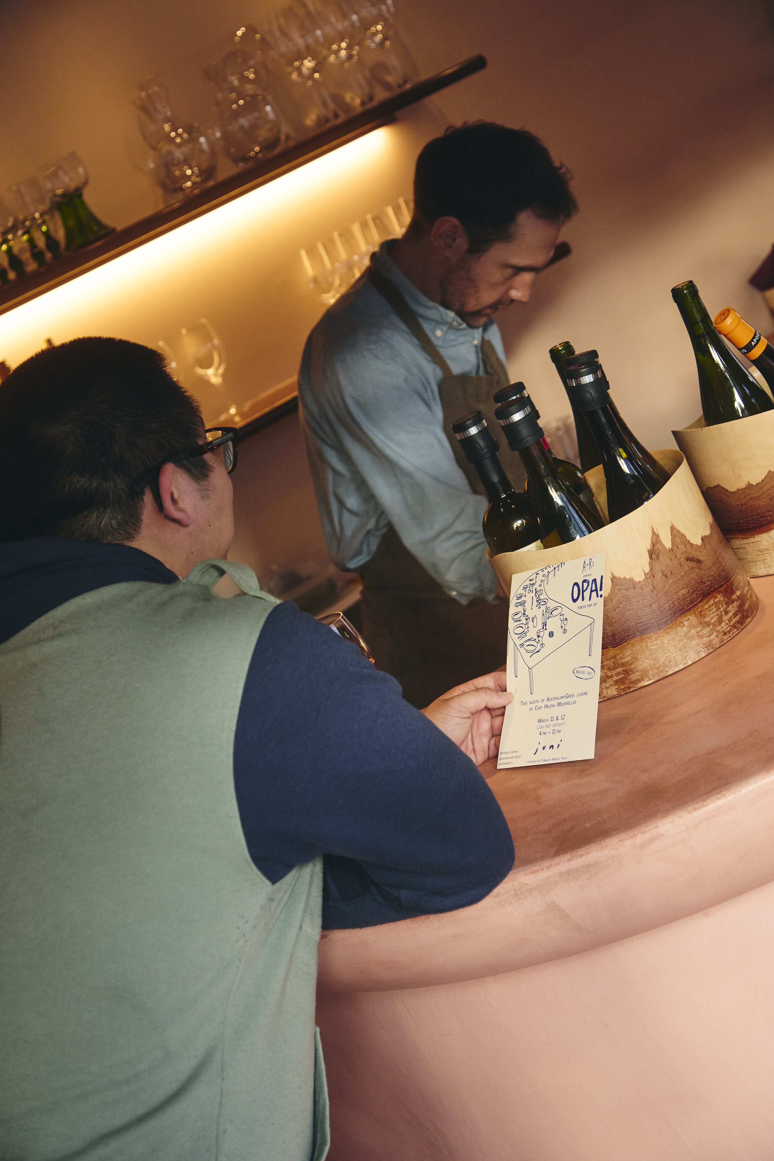A man and a woman at a bar with bottles of wine and wine glasses. The woman is holding a flyer for a dinner event.