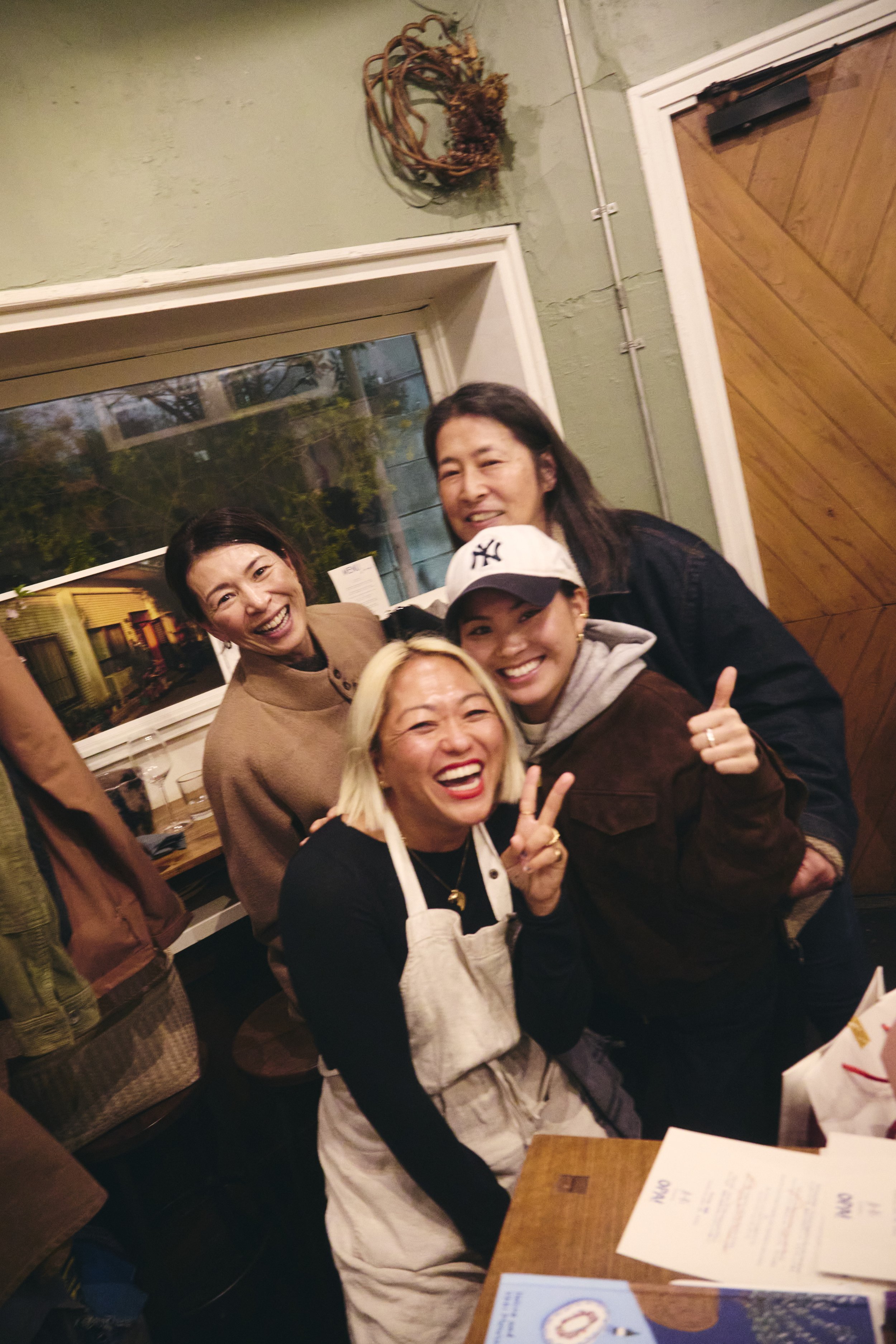 A group of smiling women and a young man posing together in a warmly lit restaurant. They are all making peace signs and look happy.