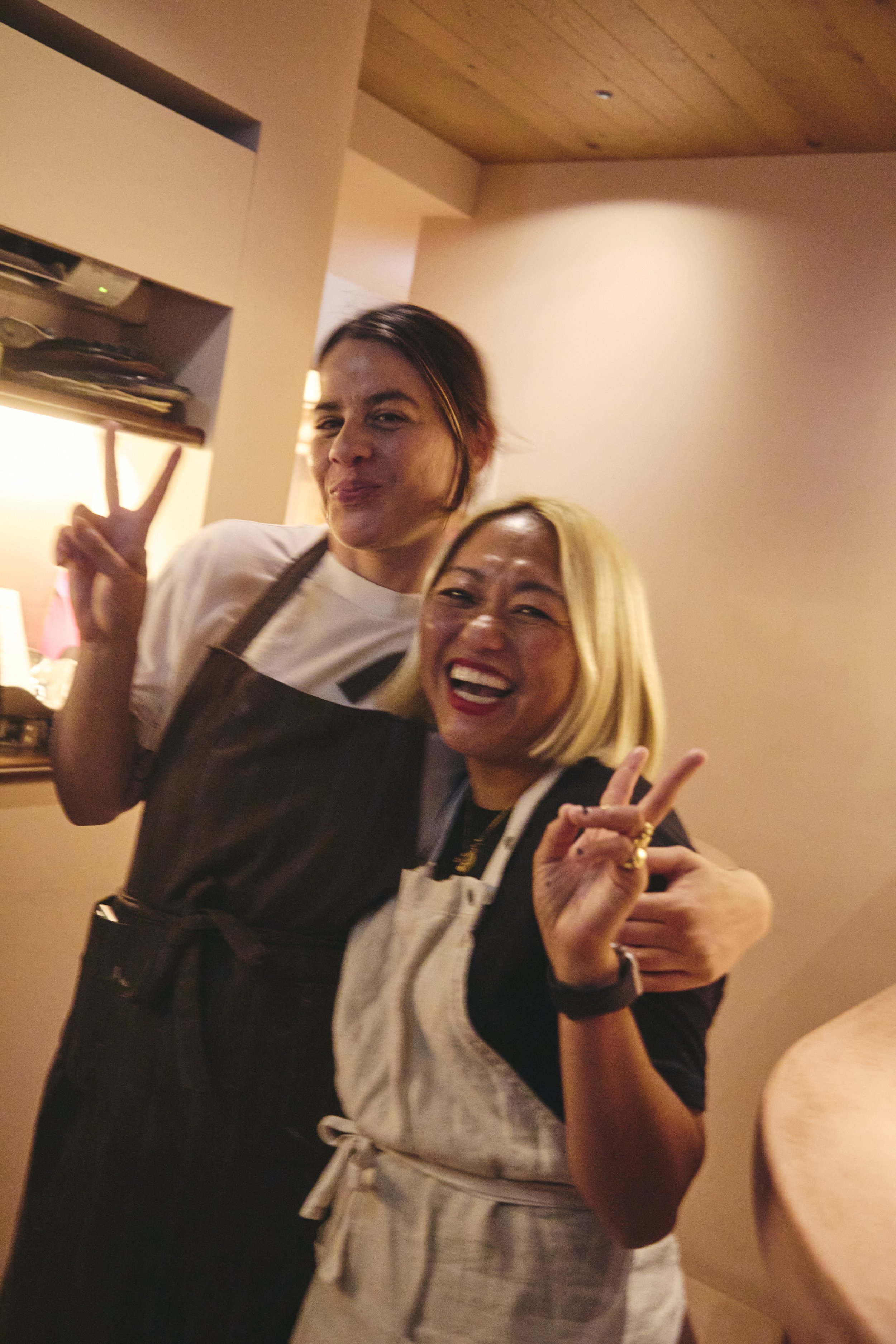 Two women posing for a photo indoors. The woman in front has blonde hair and is making a peace sign with her fingers, smiling widely. The woman behind has dark hair and is making a peace sign as well, with a slight smile. They are dressed casually, a