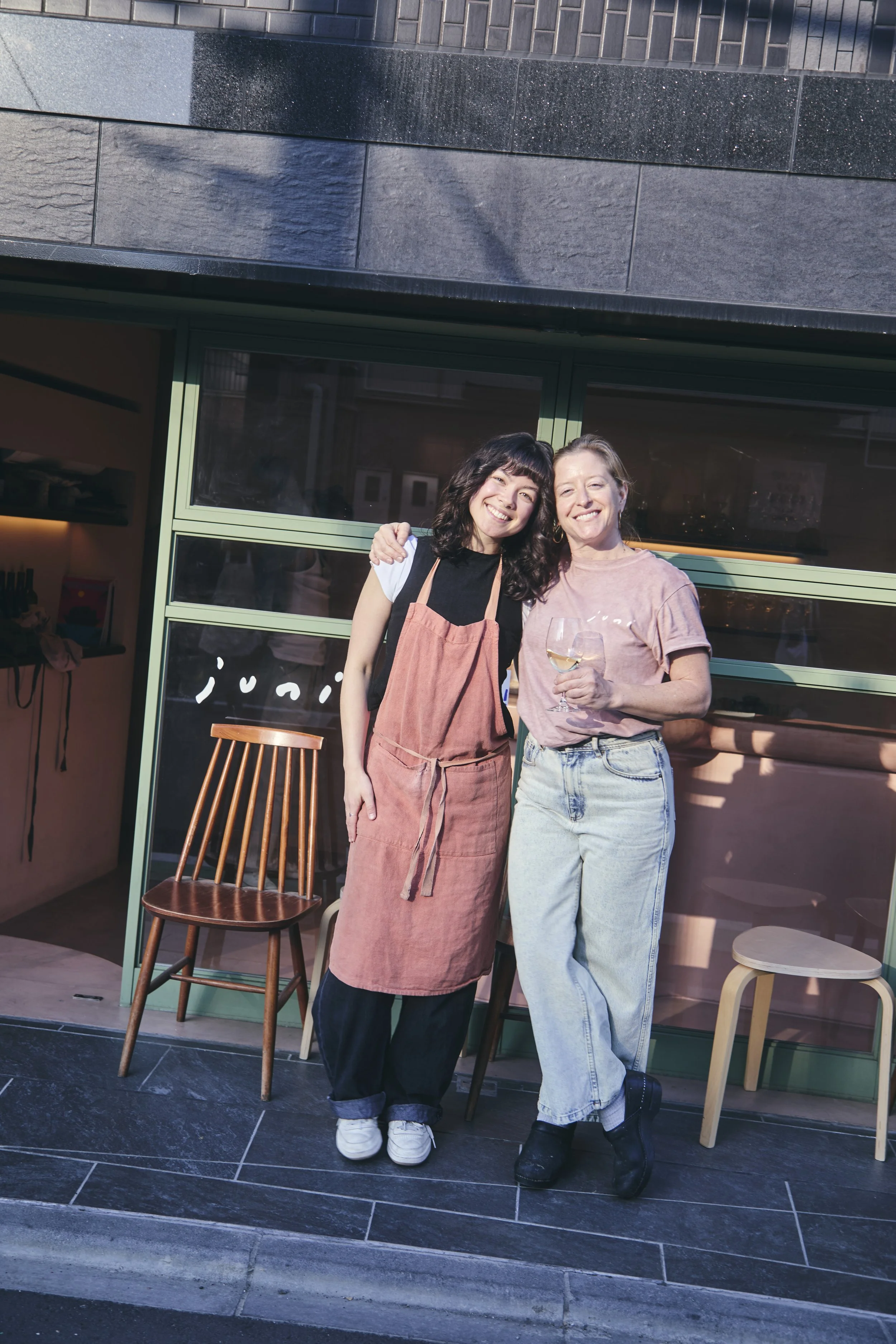 Two women standing outside a building, smiling and embracing. One holds a glass of white wine. The woman on the left wears a black T-shirt, pink apron, and black pants. The woman on the right wears a pink T-shirt and light blue jeans. There are chair