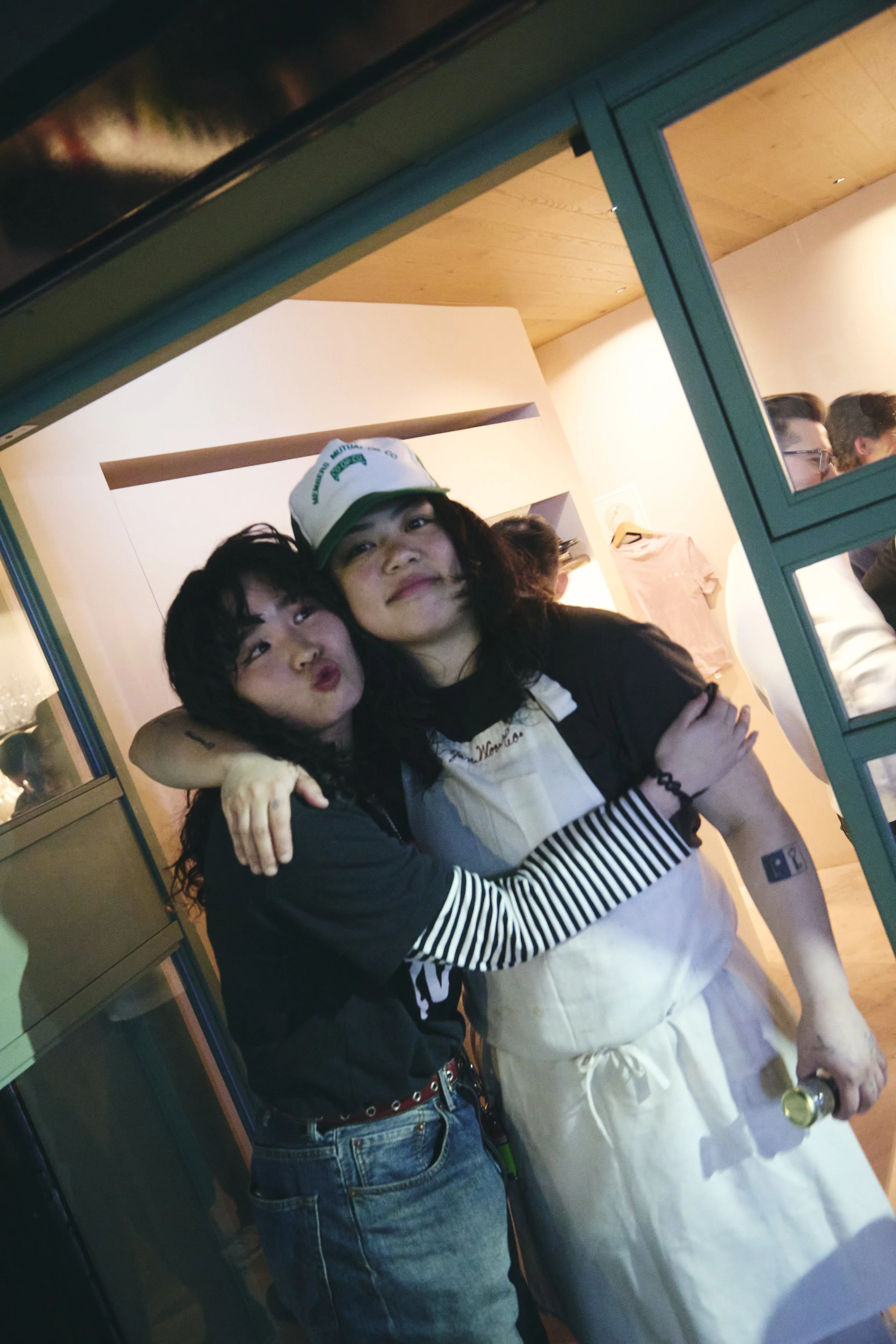 Two women hugging, one is wearing a white apron and holding a drink, the other is wearing a black sweater with white stripes and a red belt. They are inside a building, with a group of people in the background.