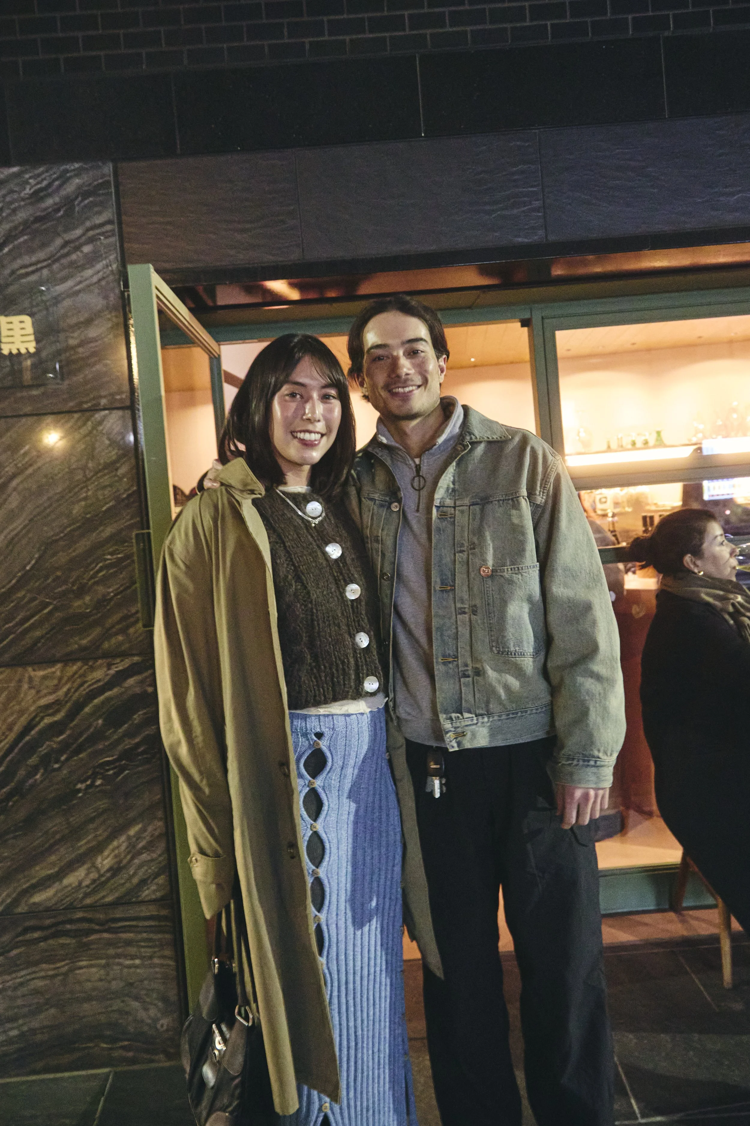 A smiling Asian woman and a smiling Caucasian man standing together indoors at night, in front of a large window with warm lighting.