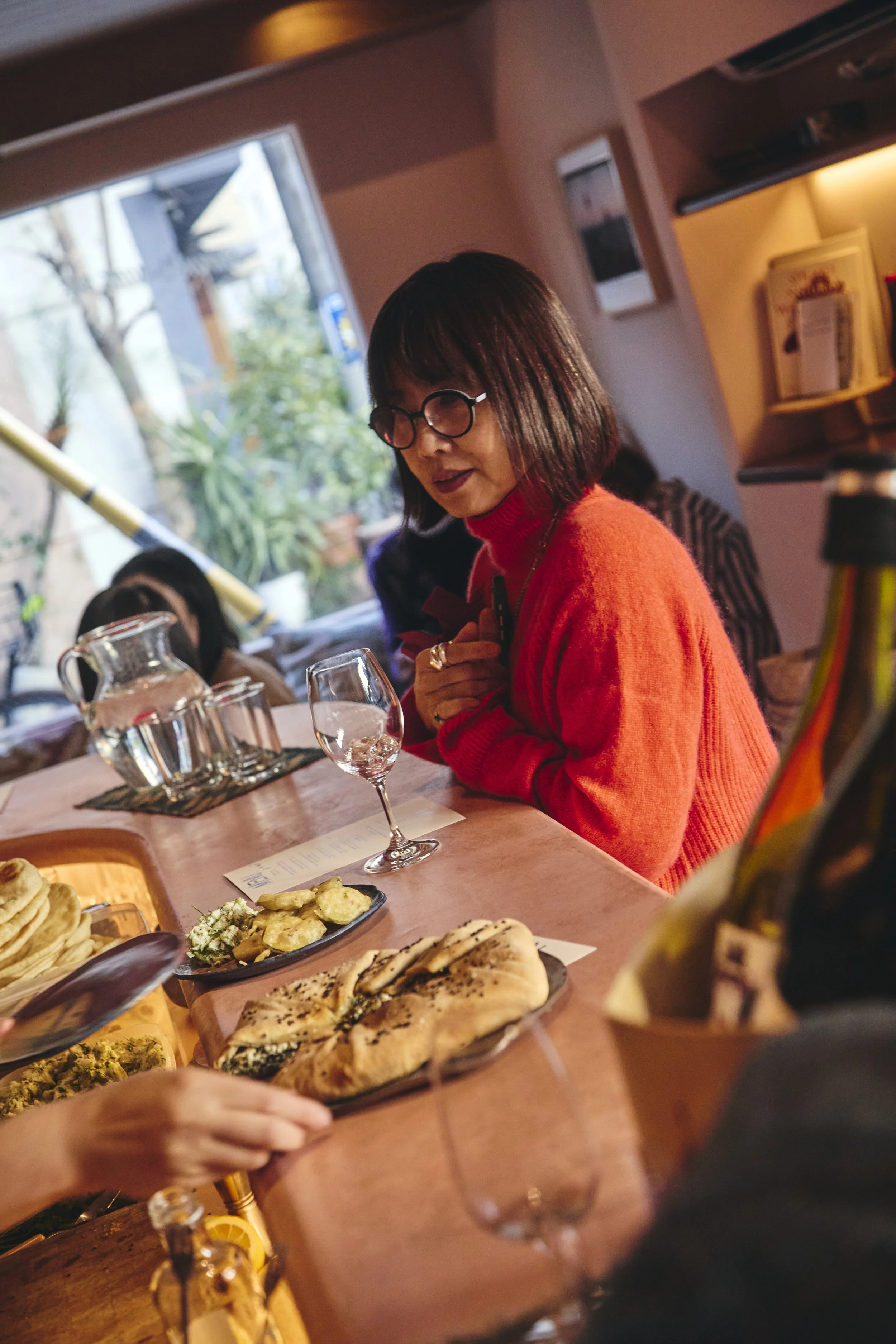 A woman with dark hair, glasses, and a red sweater sitting at a dining table, surrounded by plates of food and drinks in a cozy, warmly lit restaurant with a window view of trees outside.