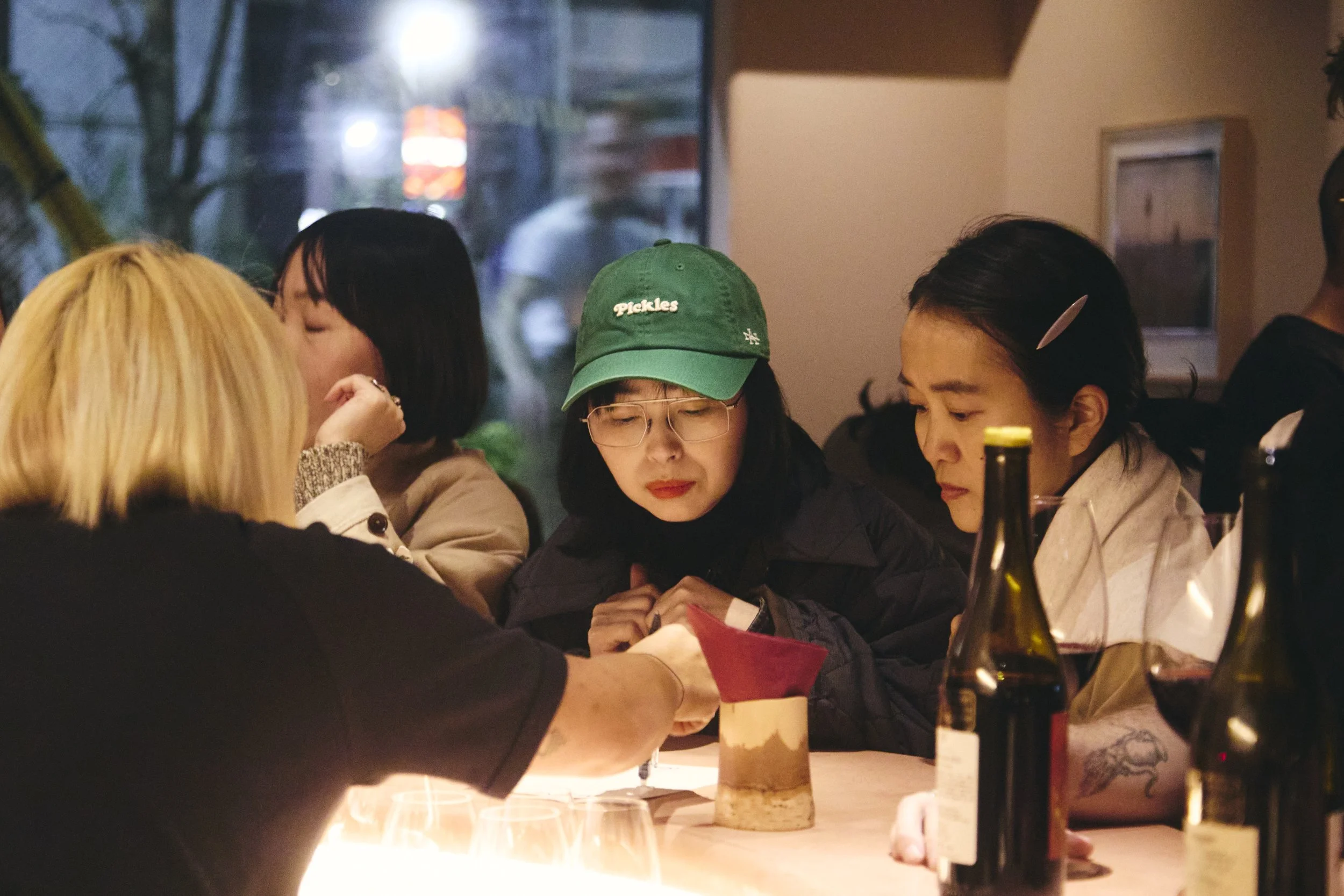A group of people gathered around a table in a dimly lit restaurant, with a woman wearing glasses and a green cap among them. There are wine bottles on the table and blurred lights visible through the window in the background.