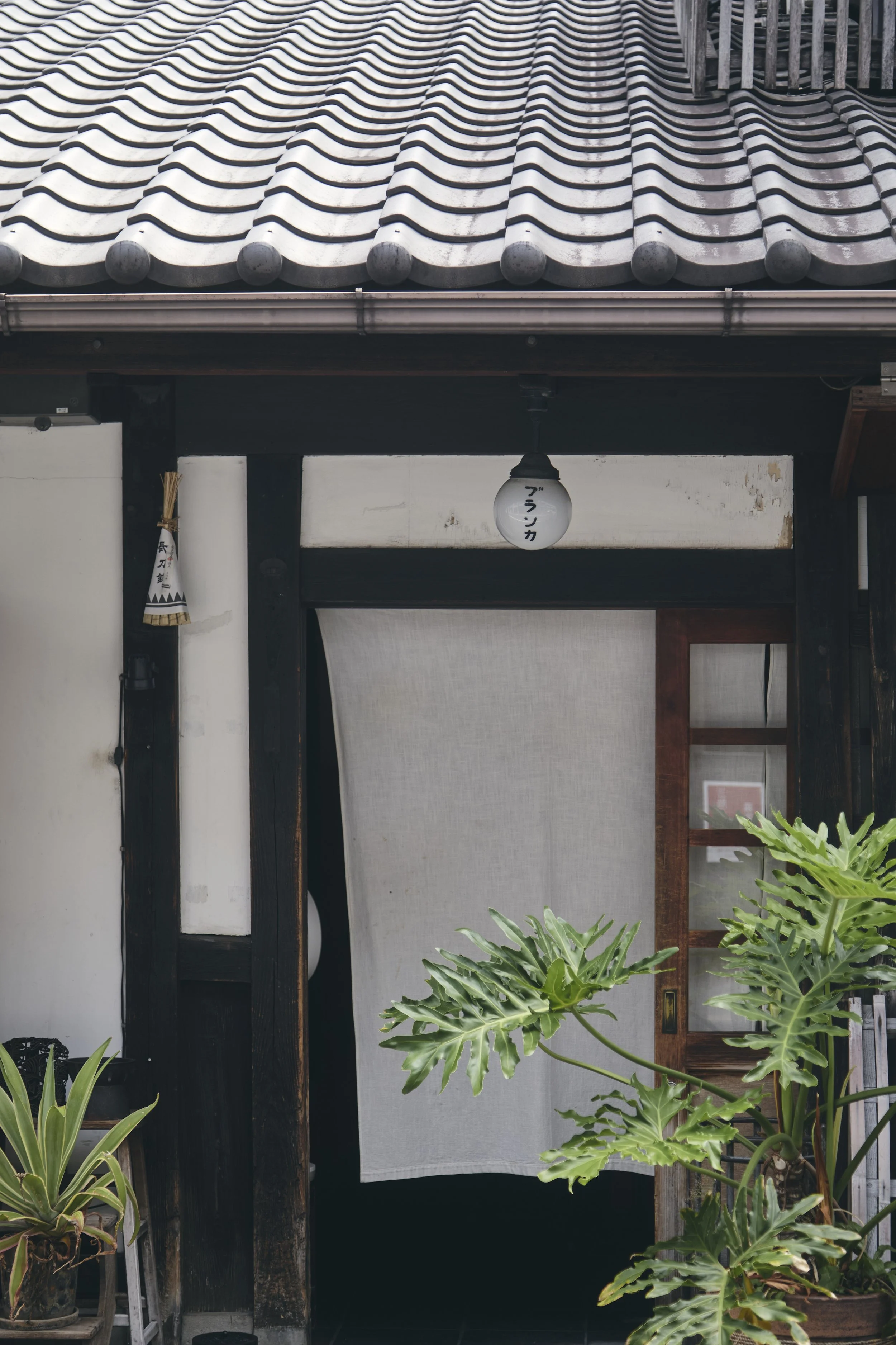 Traditional Japanese building entrance with tiled roof, wooden door, potted plants, and a paper lantern with Japanese characters.