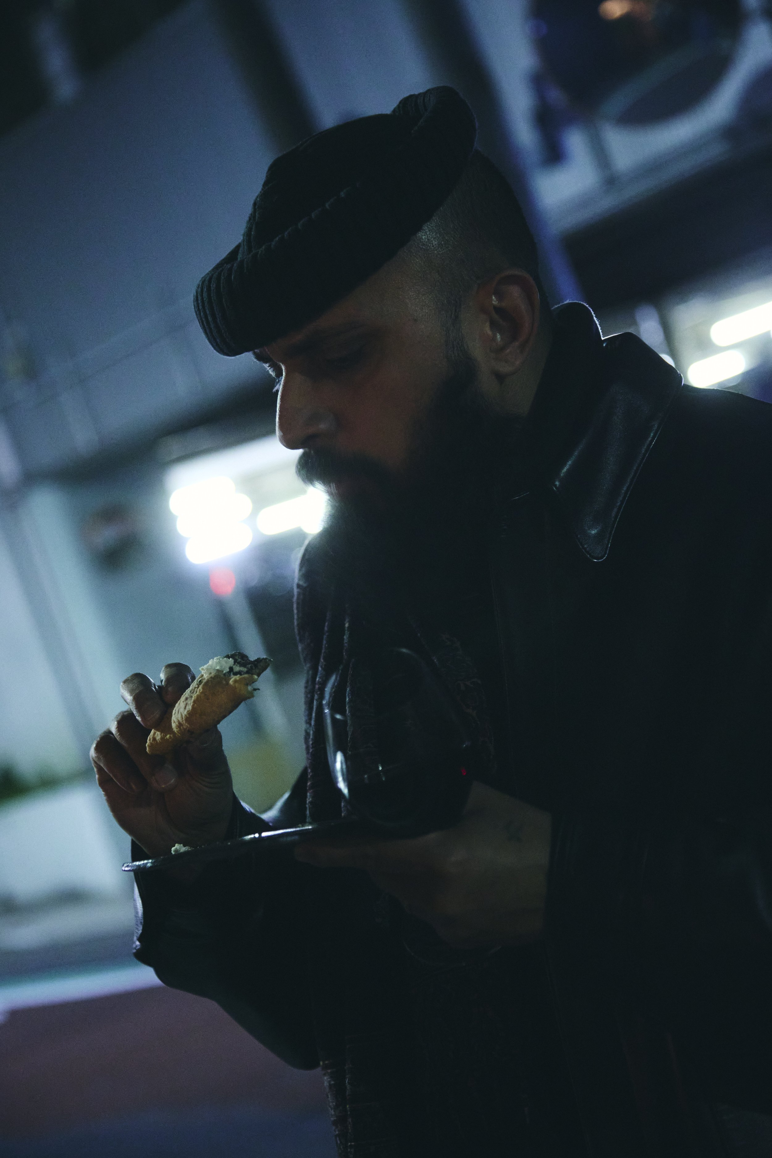 A man with a beard, wearing a black hat and black leather jacket, is holding a plate with a sandwich, eating it in a dimly lit indoor setting with blurred background lights.