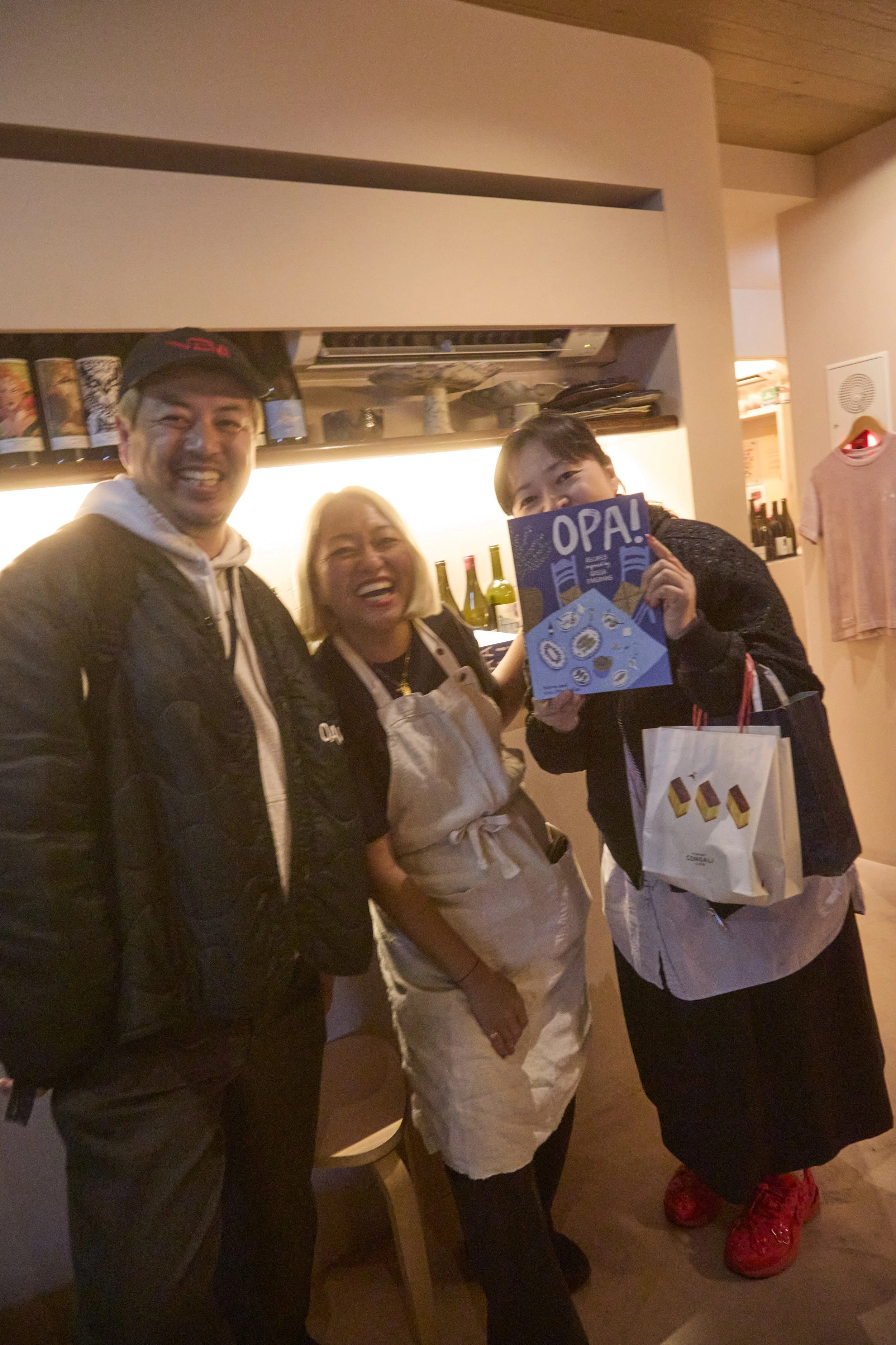 Three people, two women and one man, smiling and standing together indoors. One woman is holding a blue card that says 'OPA!' and a shopping bag. They seem happy and are dressed casually.