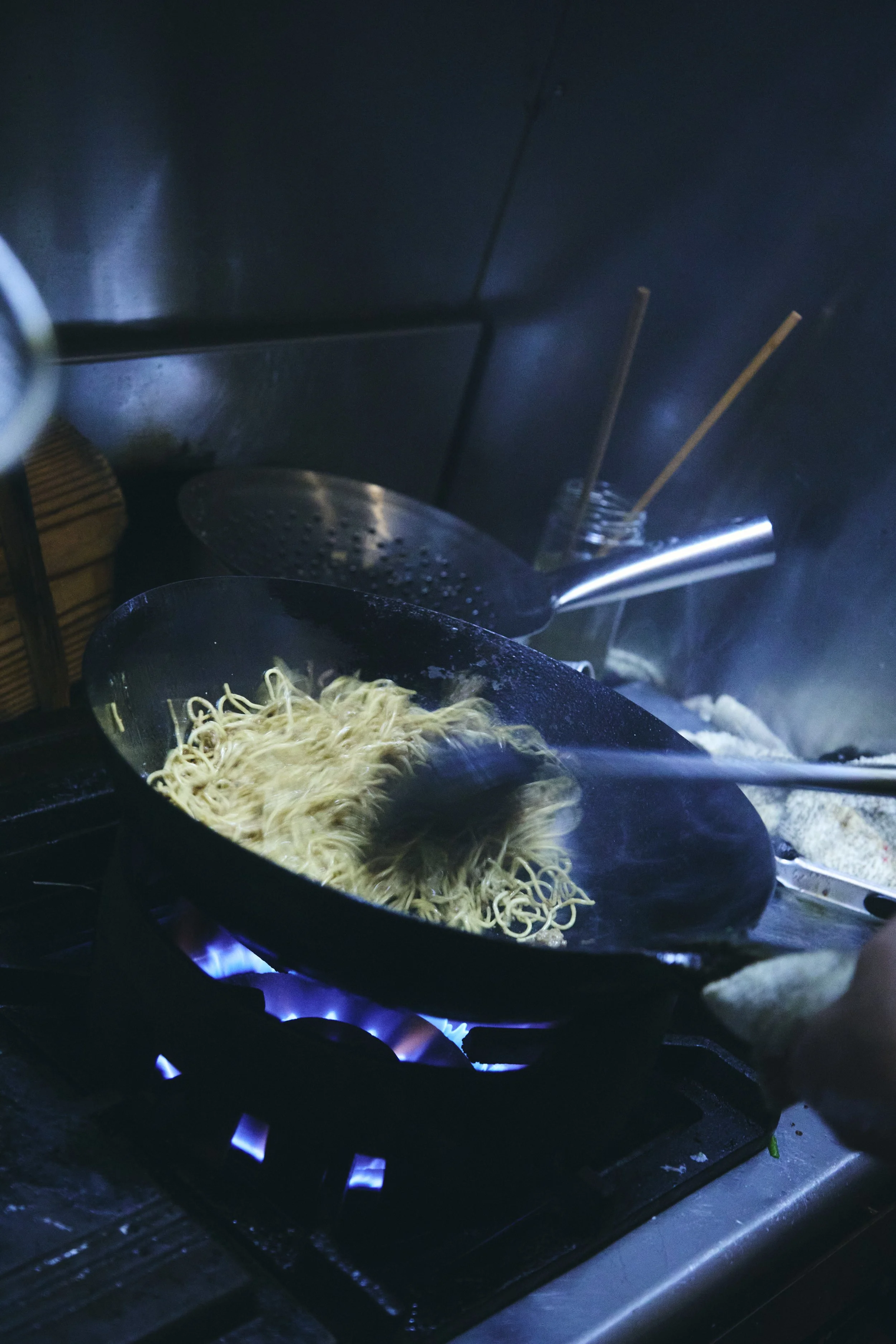 Cooking noodles in a black pan over a blue flame gas stove, with chopsticks and a spatula nearby.