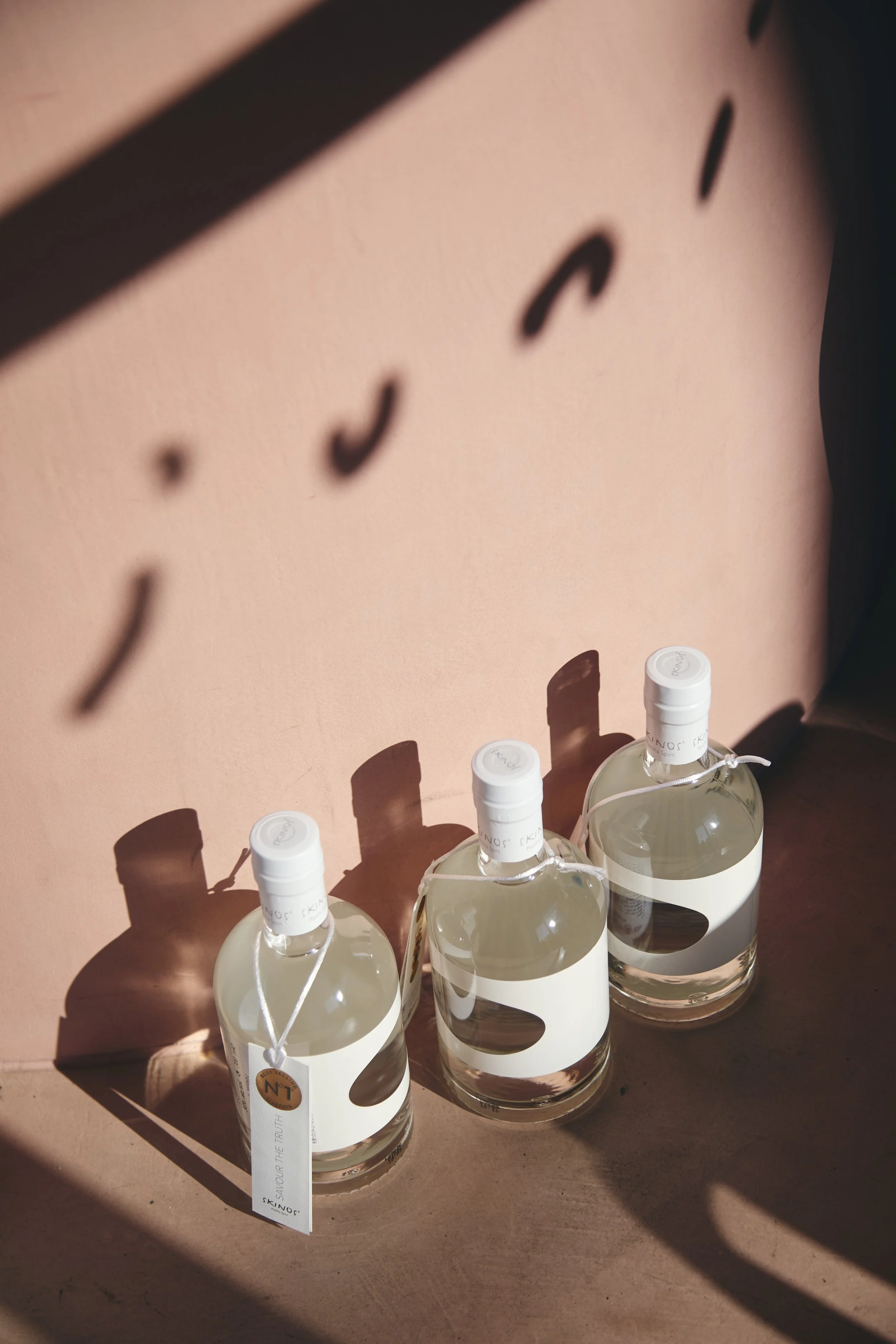 Four bottles of clear liquid with white caps and labels, placed in a wooden holder with circular cutouts. Shadows cast on a pink wall behind them.