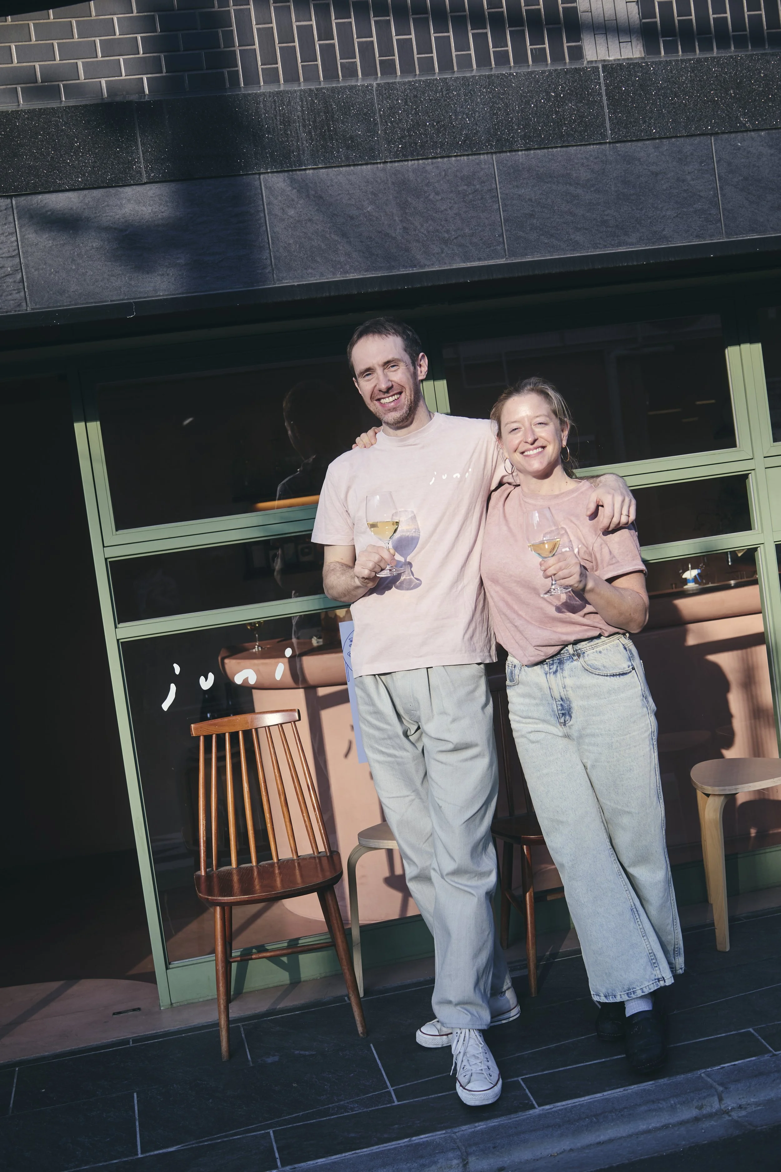 A young man and woman smiling and raising wine glasses outside a cafe or restaurant, with a green-framed window behind them