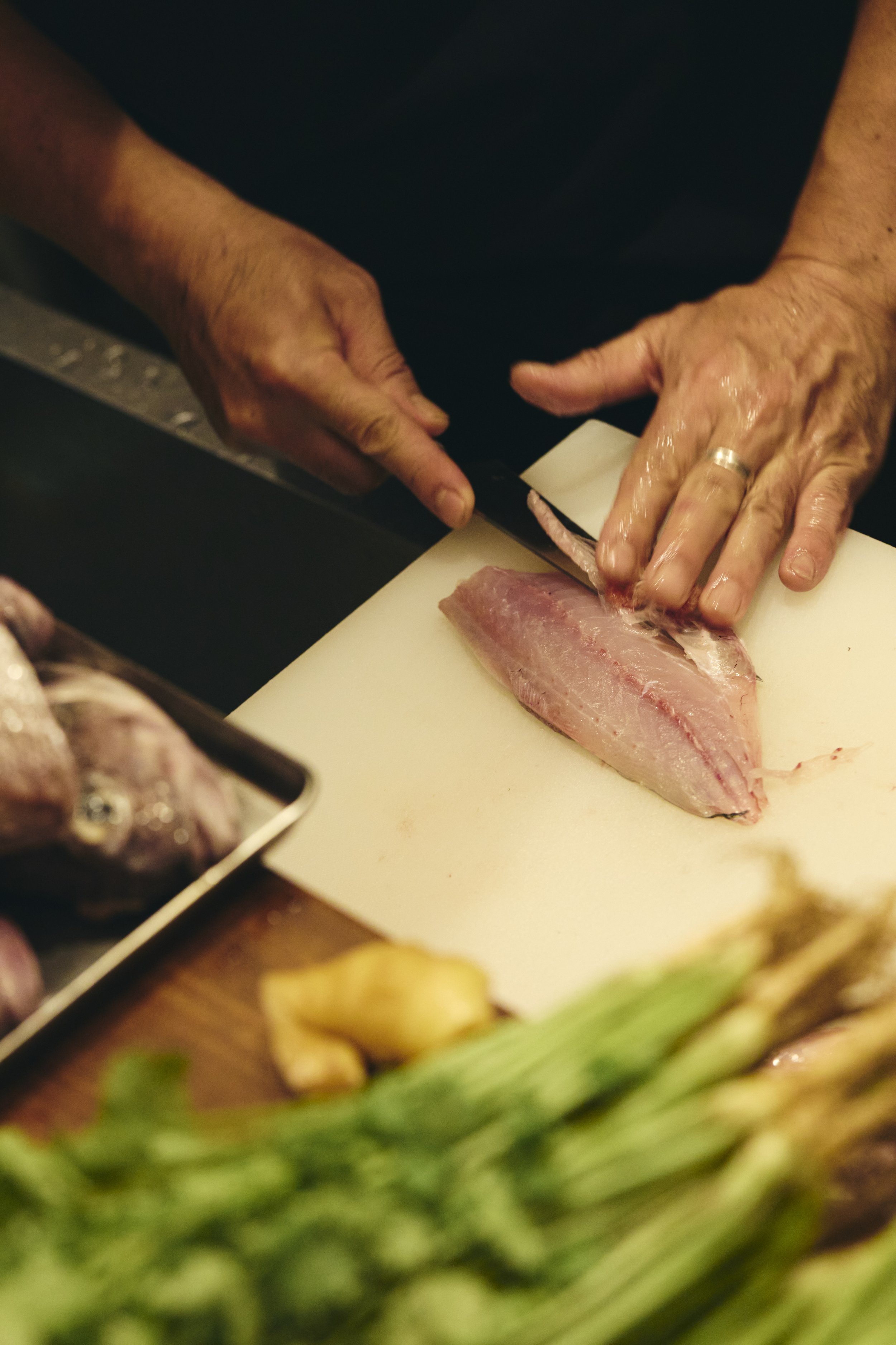 Person slicing a piece of fish on a white cutting board in a kitchen, with fresh vegetables and other ingredients in the foreground.