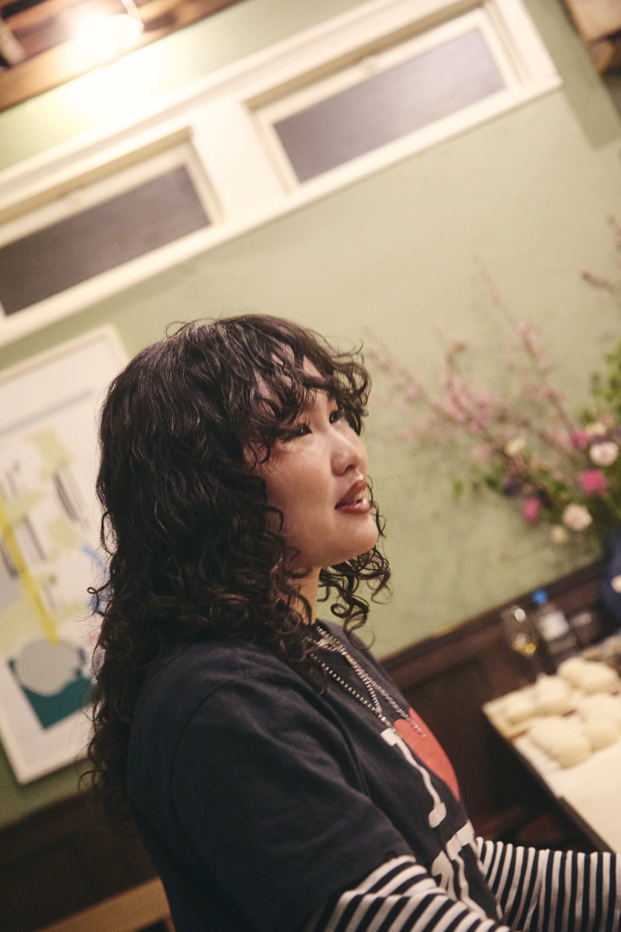 A woman with curly dark hair sitting indoors in front of a green wall with pink cherry blossom decorations, wearing a black jacket and striped shirt, looking to her left.