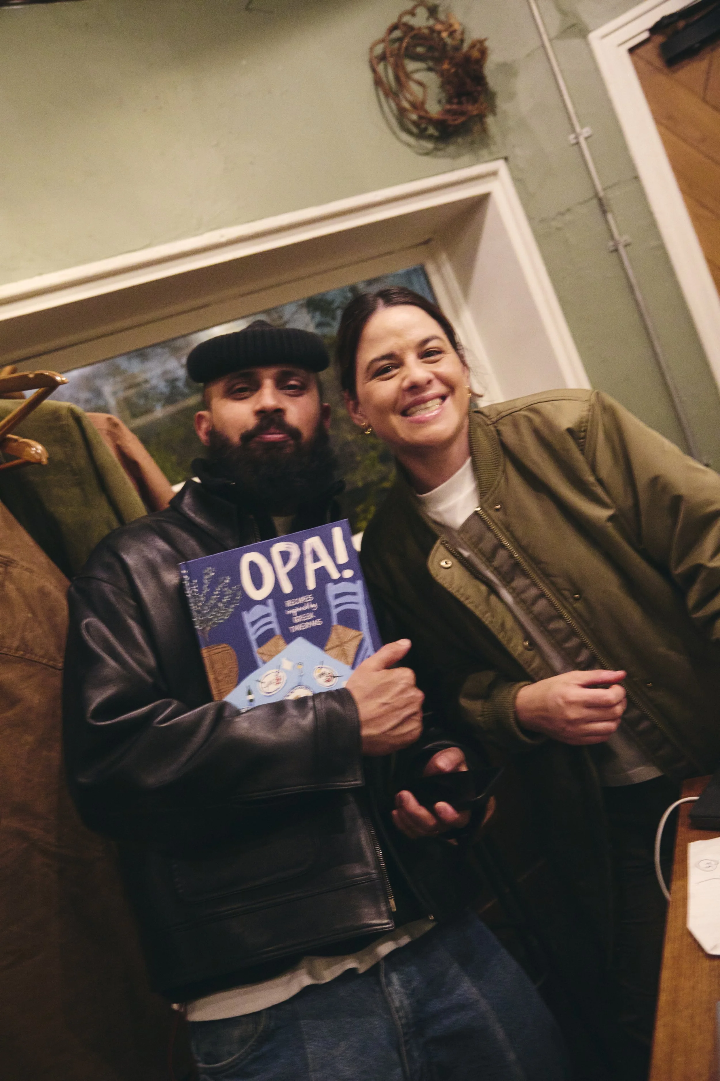A man with a beard wearing a black hat and leather jacket holding a blue book, standing next to a woman smiling and wearing a green jacket, inside a room with a green wall and a window in the background.