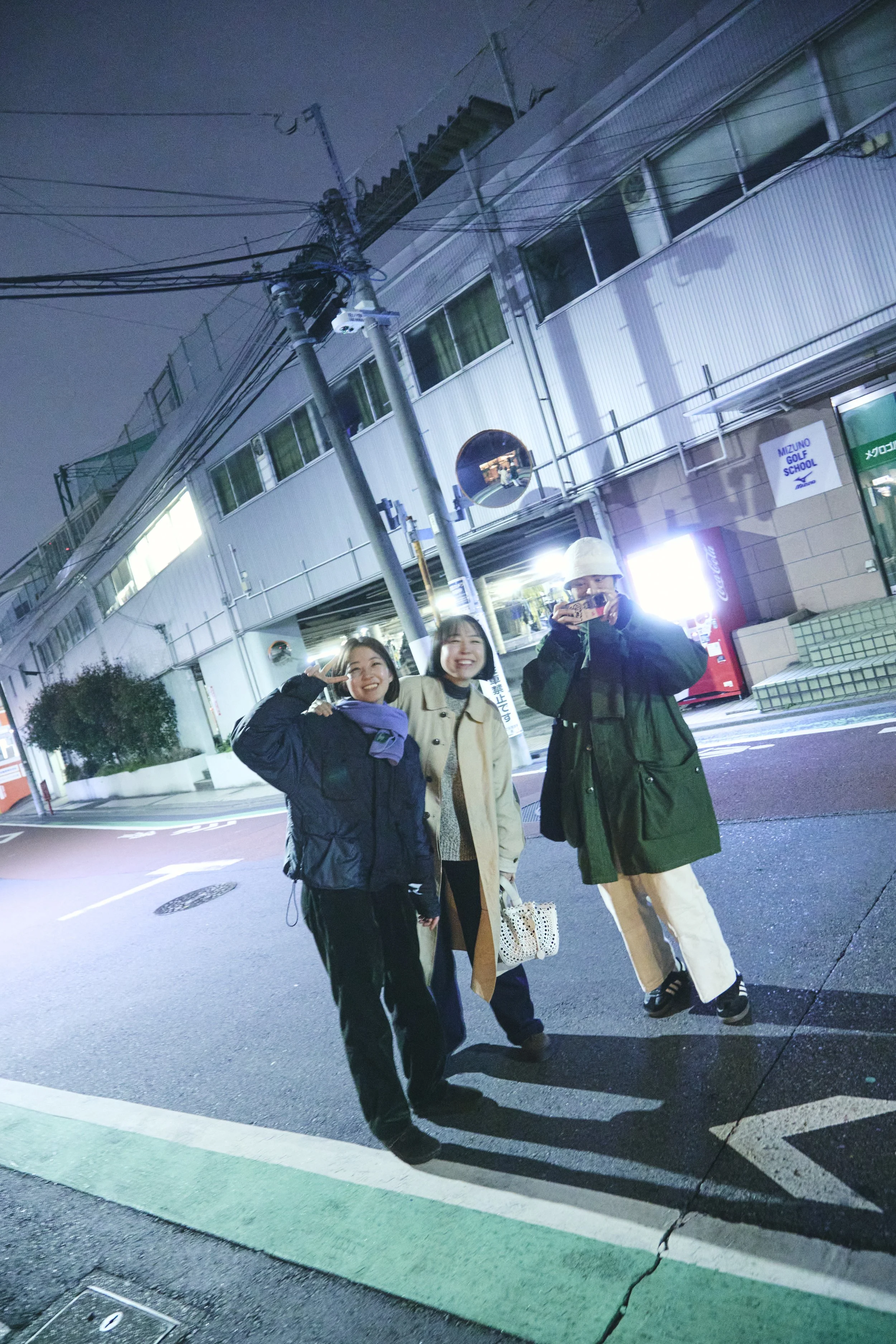 Three young people standing on a city street at night, smiling and posing for the photo. They are wearing jackets, and the background features a modern building with signs for Mizuno Golf School and a Coca-Cola vending machine.