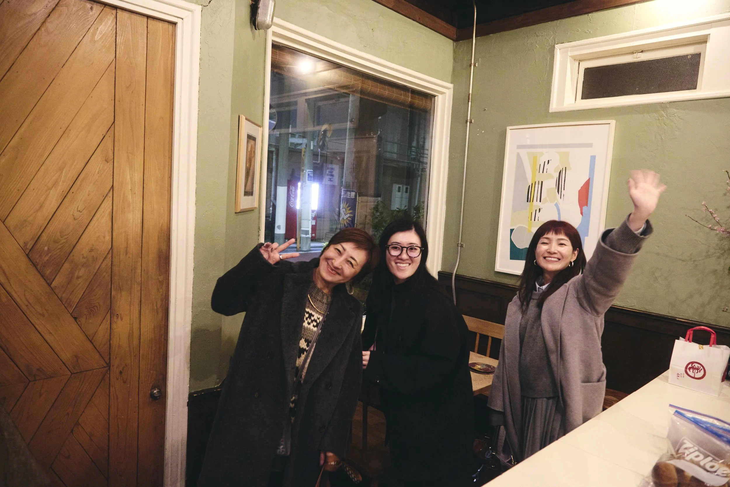 Three women smiling and posing for a photo inside a restaurant. One woman on the right is raising her hand, and the other two are standing close together, making peace signs.