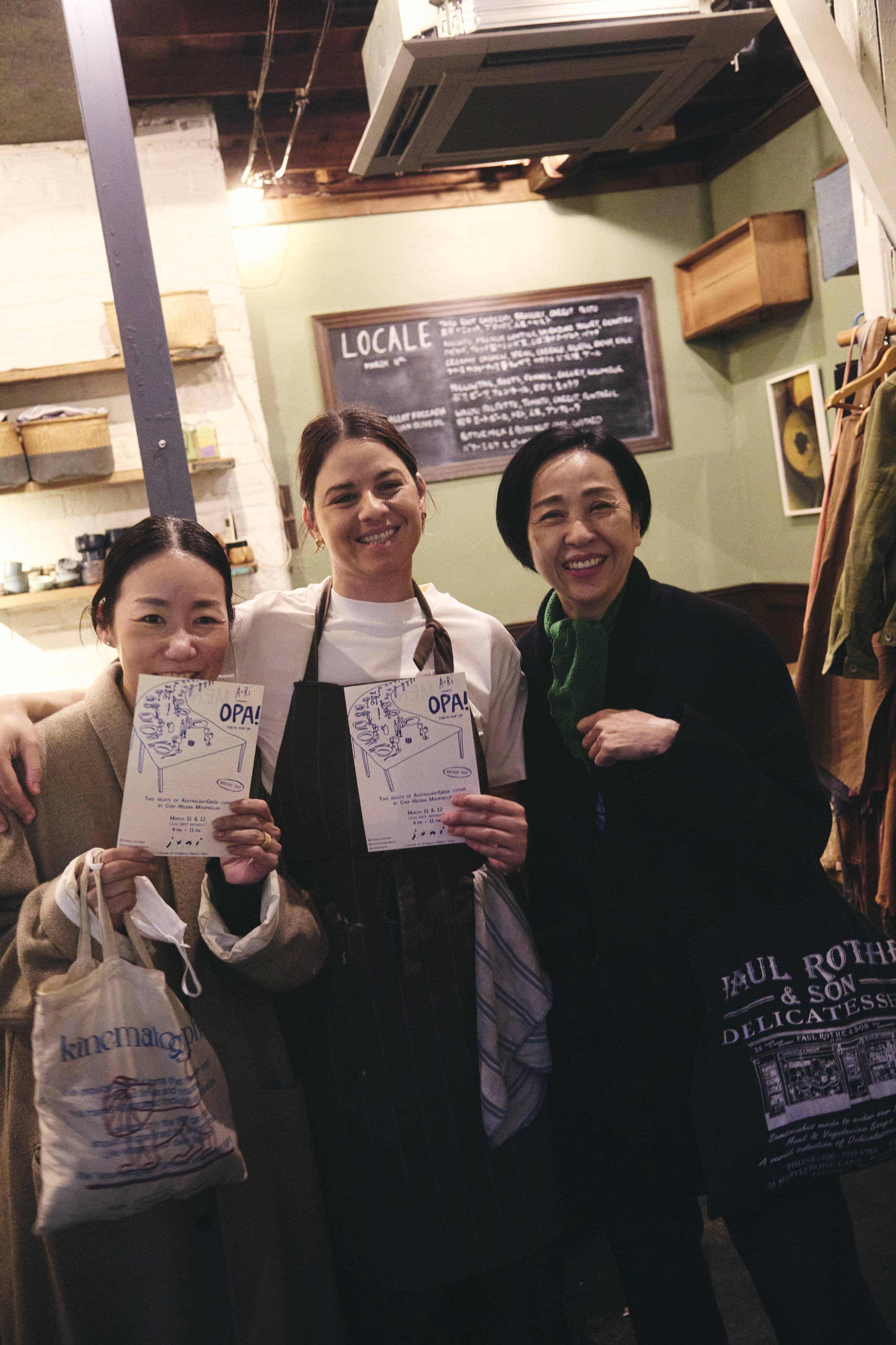 Three women smiling and holding papers, posing inside a cozy restaurant. A chalkboard menu is visible on the wall behind them, and shelves with baskets and other items are seen on the left side.