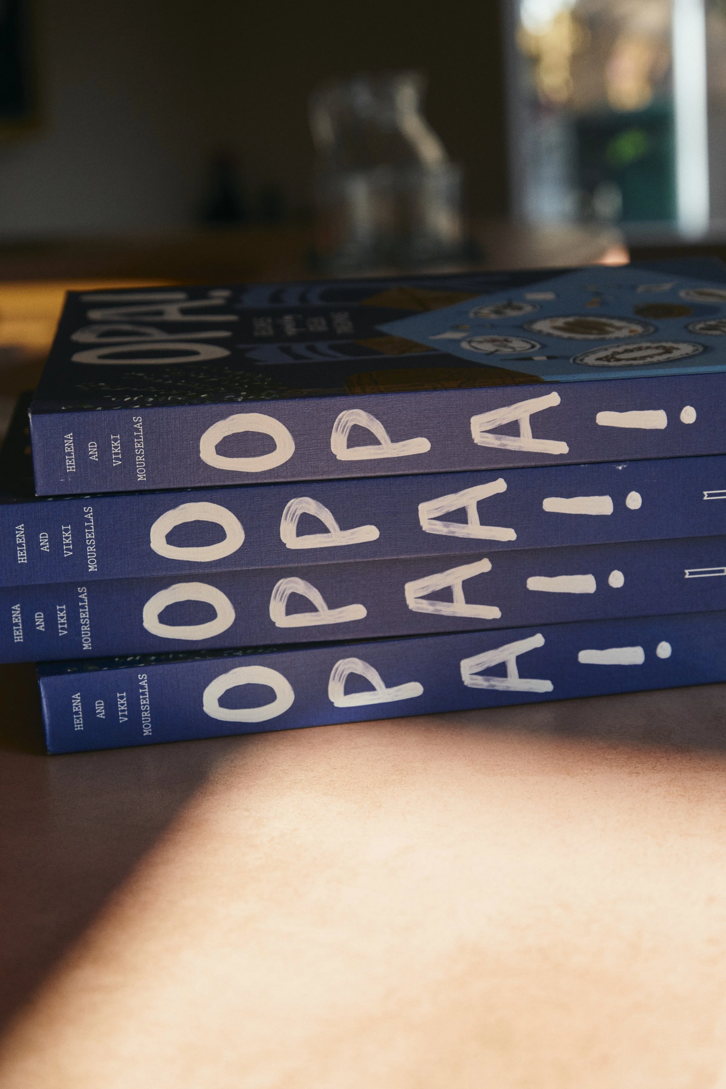Close-up of a stack of three copies of a book titled "PADDLE" by Helena Vikklas, with the titles visible on the spines. The books are placed on a surface with sunlight and a blurred background.