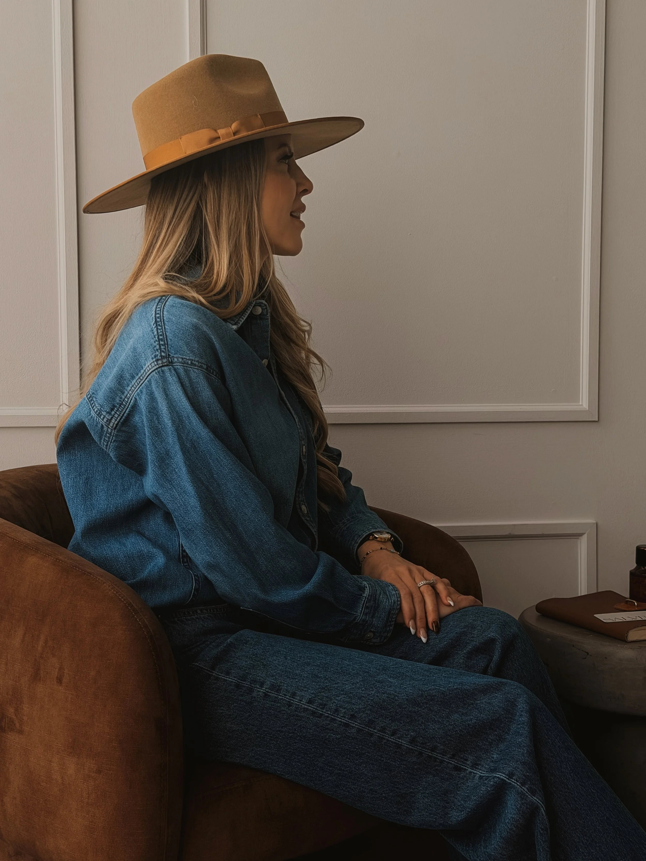 A woman with long, wavy hair wearing a wide-brimmed tan hat, denim jacket, and jeans is sitting in profile on a brown couch in a room with beige walls and white paneling.