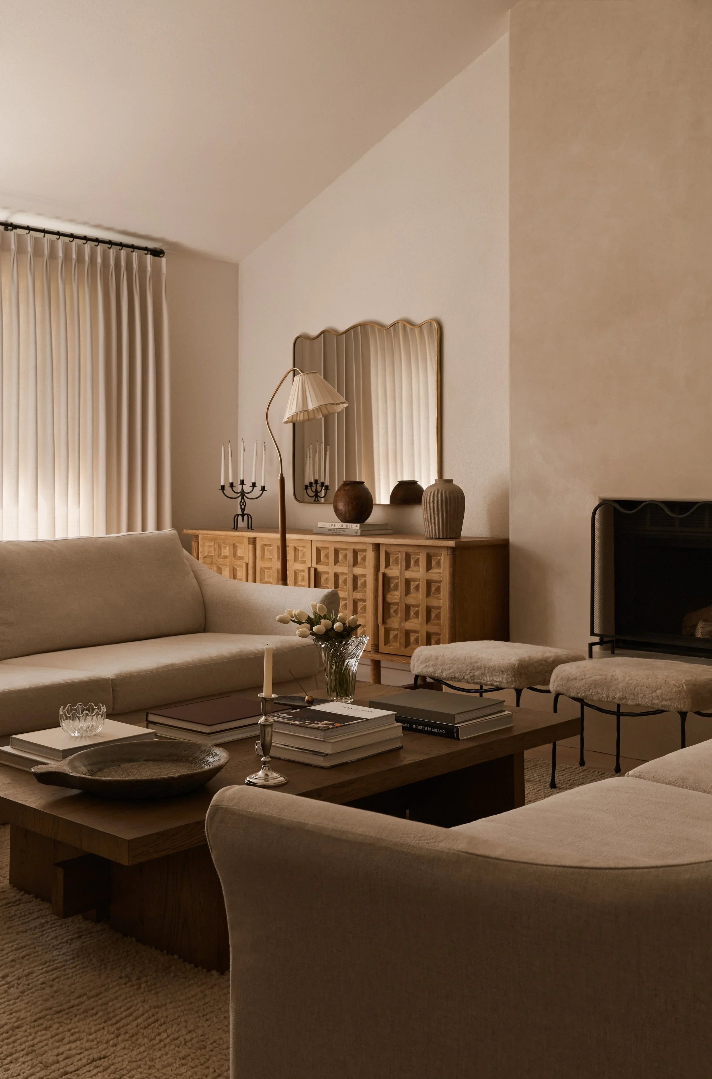 A cozy living room with beige sofas, a wooden coffee table with books and a candle, and a sideboard with vases, a mirror, and a lamp. Cream curtains cover the window, and a beige carpet completes the warm neutral decor.