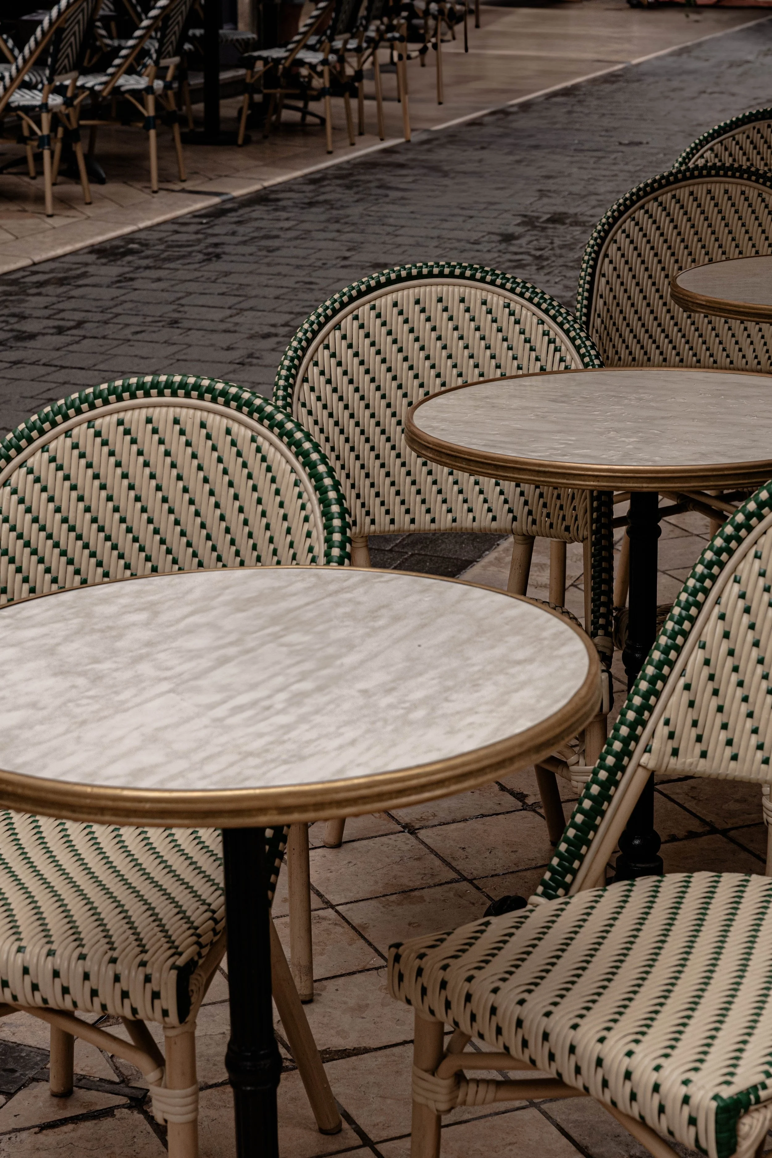 Empty outdoor cafe seating with beige and green woven chairs, round marble-topped tables, and brick patio flooring.