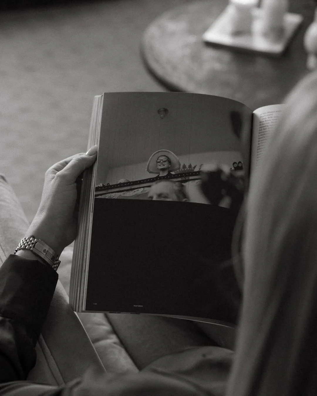 A person holding a magazine or book, showing a black-and-white photo of a woman wearing glasses and a wide-brimmed hat, smiling outdoors.