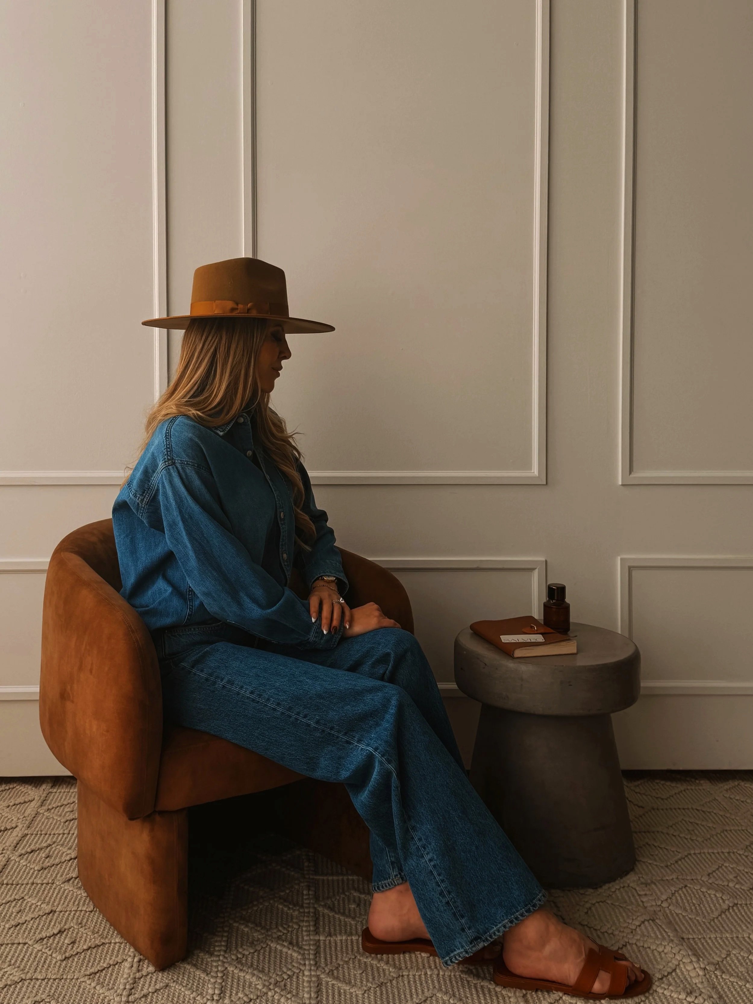 A woman with long hair wearing a wide-brimmed hat, denim jacket, and matching jeans sits on an orange armchair in a room with beige walls. She is facing sideways with her hands resting on her lap. There is a small round table next to her with a book, a bottle, and a note. The floor is carpeted.