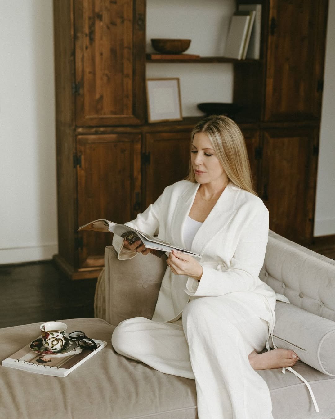A woman in white pajamas sitting on a beige sofa, reading a magazine. She is next to a coffee table with a coffee cup and magazine. Behind her is a wooden cabinet with decorative items.