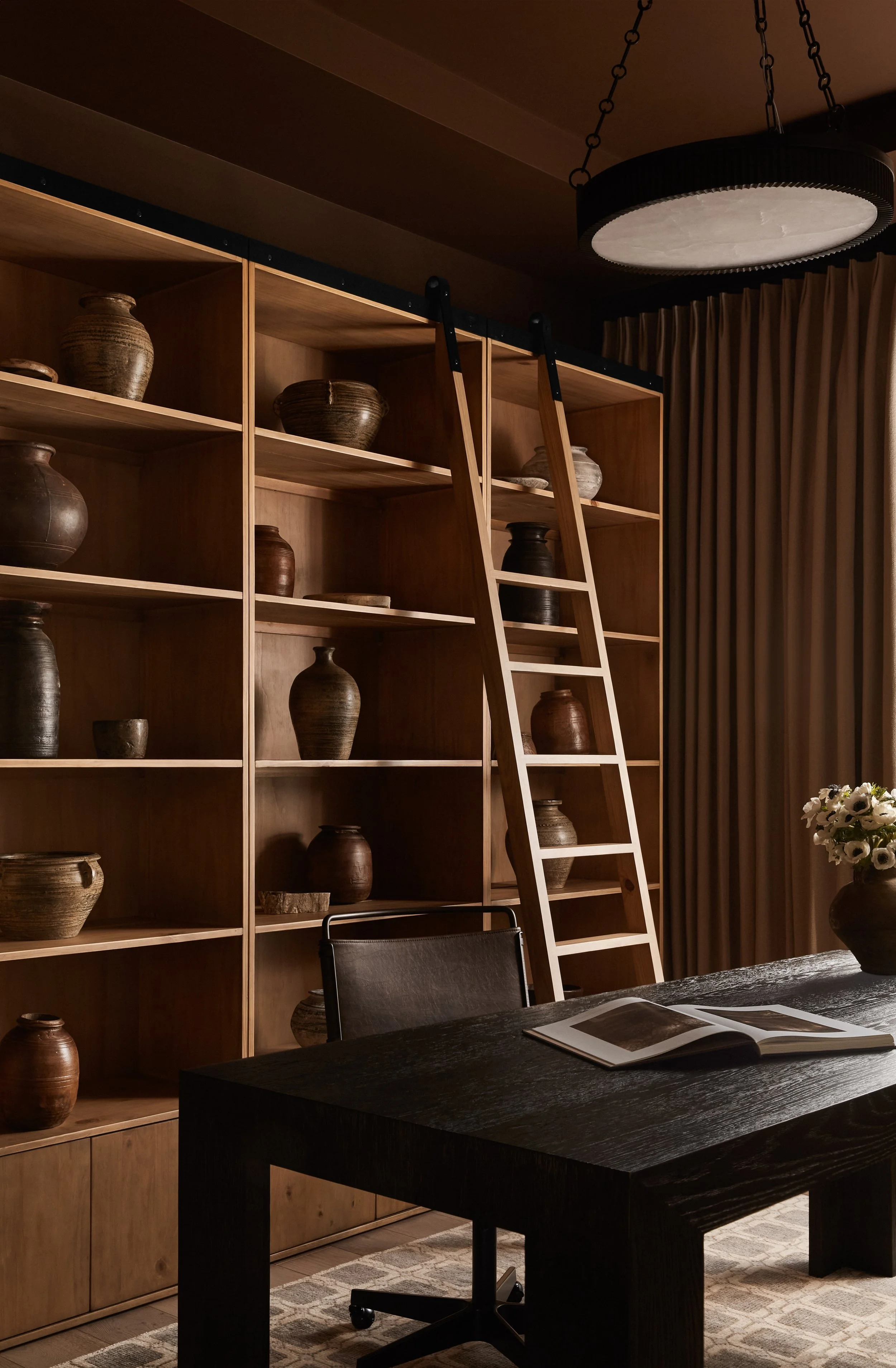 A room with wooden shelving filled with various ceramic vases, a black wooden table with an open book, black chairs, a circular ceiling light, and beige curtains.