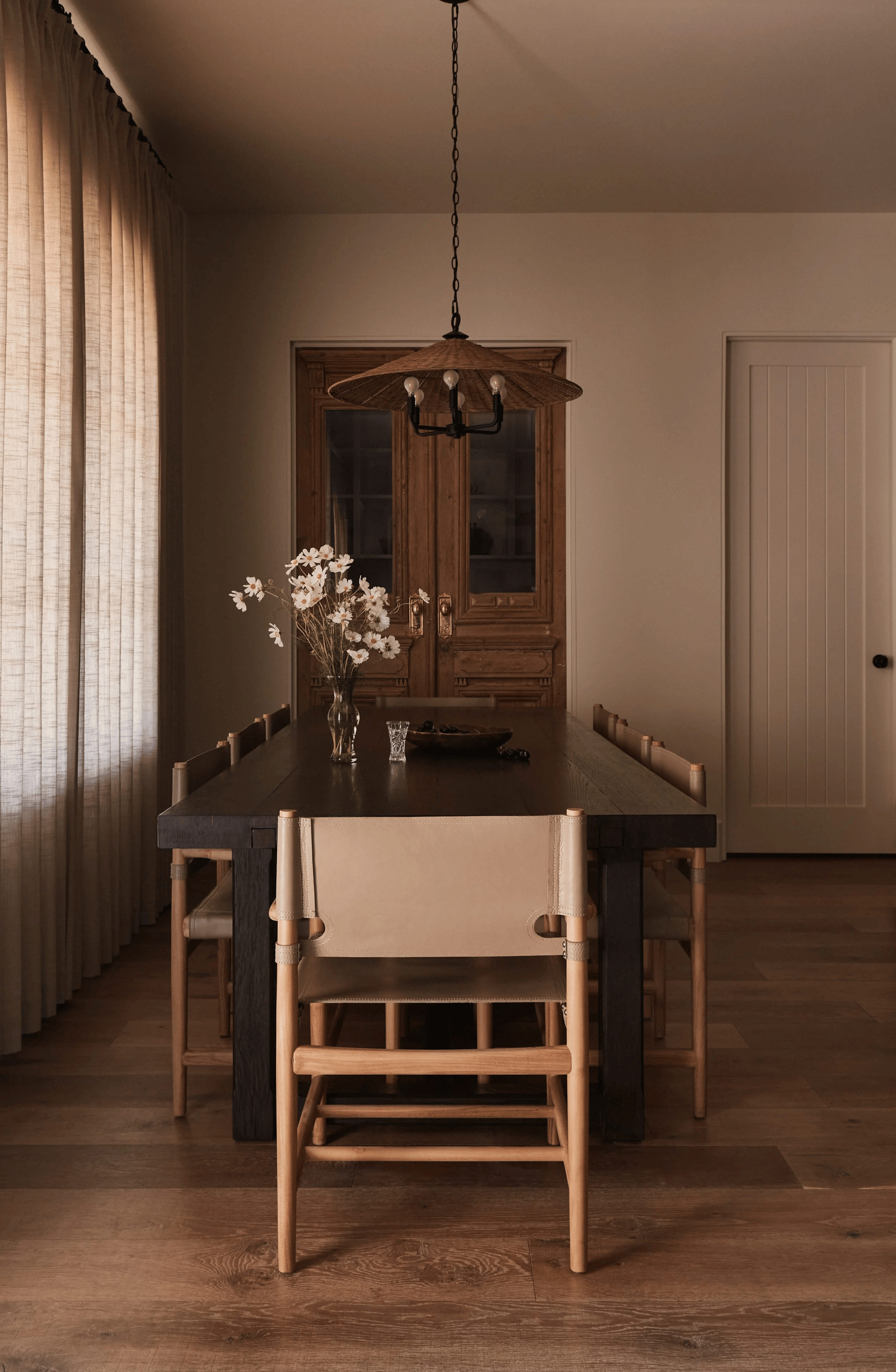 A dining room with a long dark wooden table, surrounded by wooden chairs with beige fabric seats. On the table is a glass vase with white daisies and a small glass. The room features beige curtains on the left wall, a wooden cabinet with glass doors in the background, and a chandelier with exposed bulbs hanging above the table. The flooring is wooden, and there's a white paneled door on the right side.