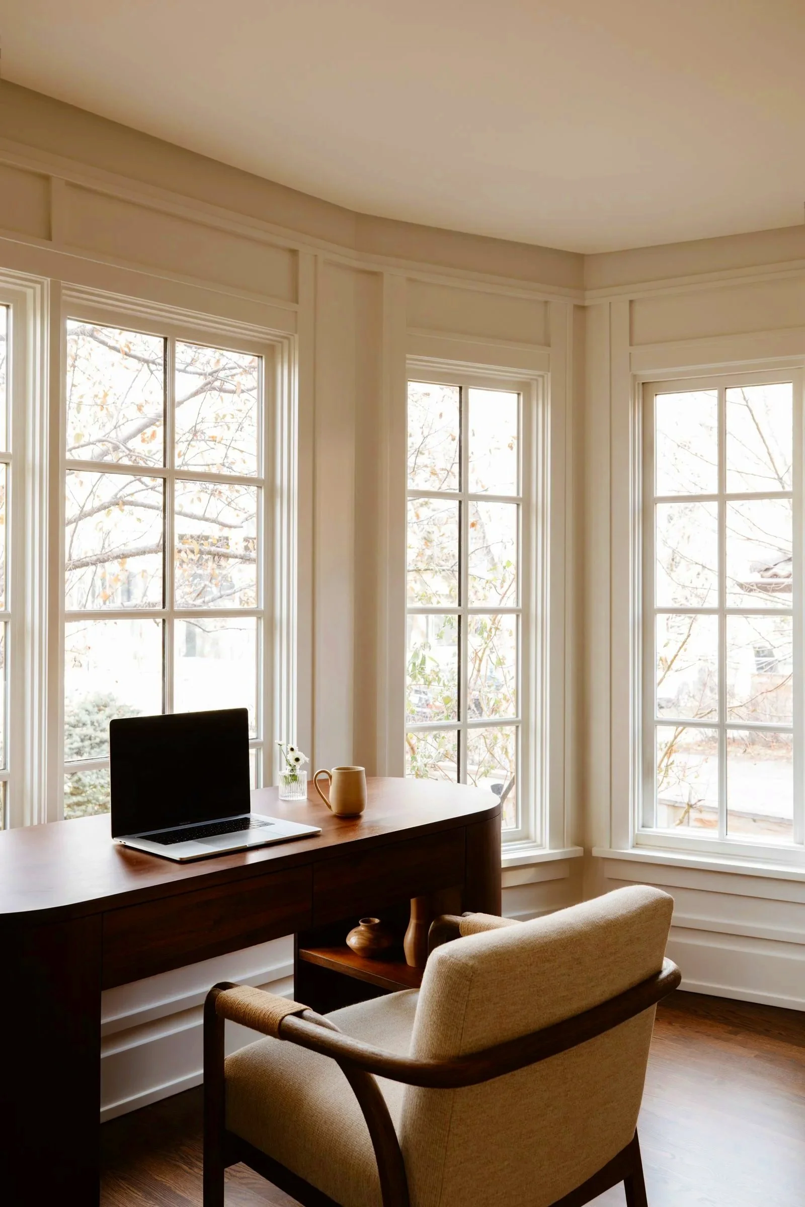 Interior of a room with large bay windows, a wooden desk with a laptop, a mug, and flowers, and a beige armchair with wooden armrests. Outside the windows, trees with fall leaves are visible.