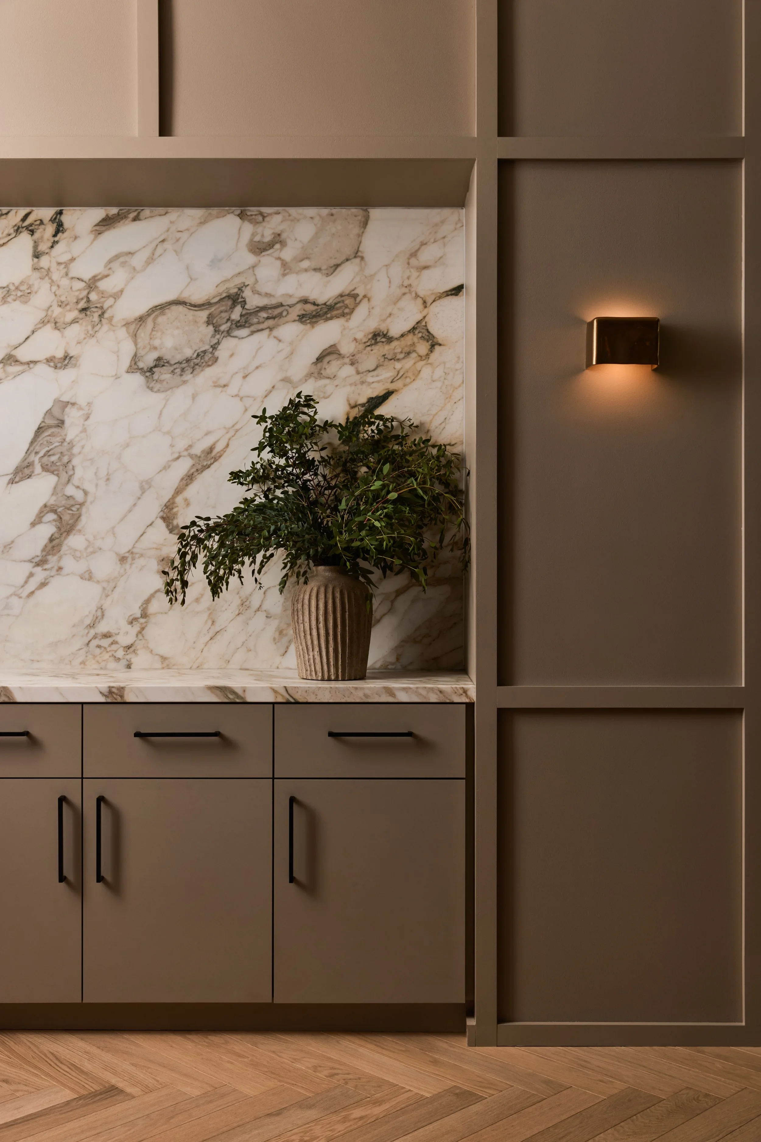 Modern kitchen cabinetry with beige doors, a marble backsplash, a potted plant in a textured beige vase, and a warm wall-mounted light fixture.