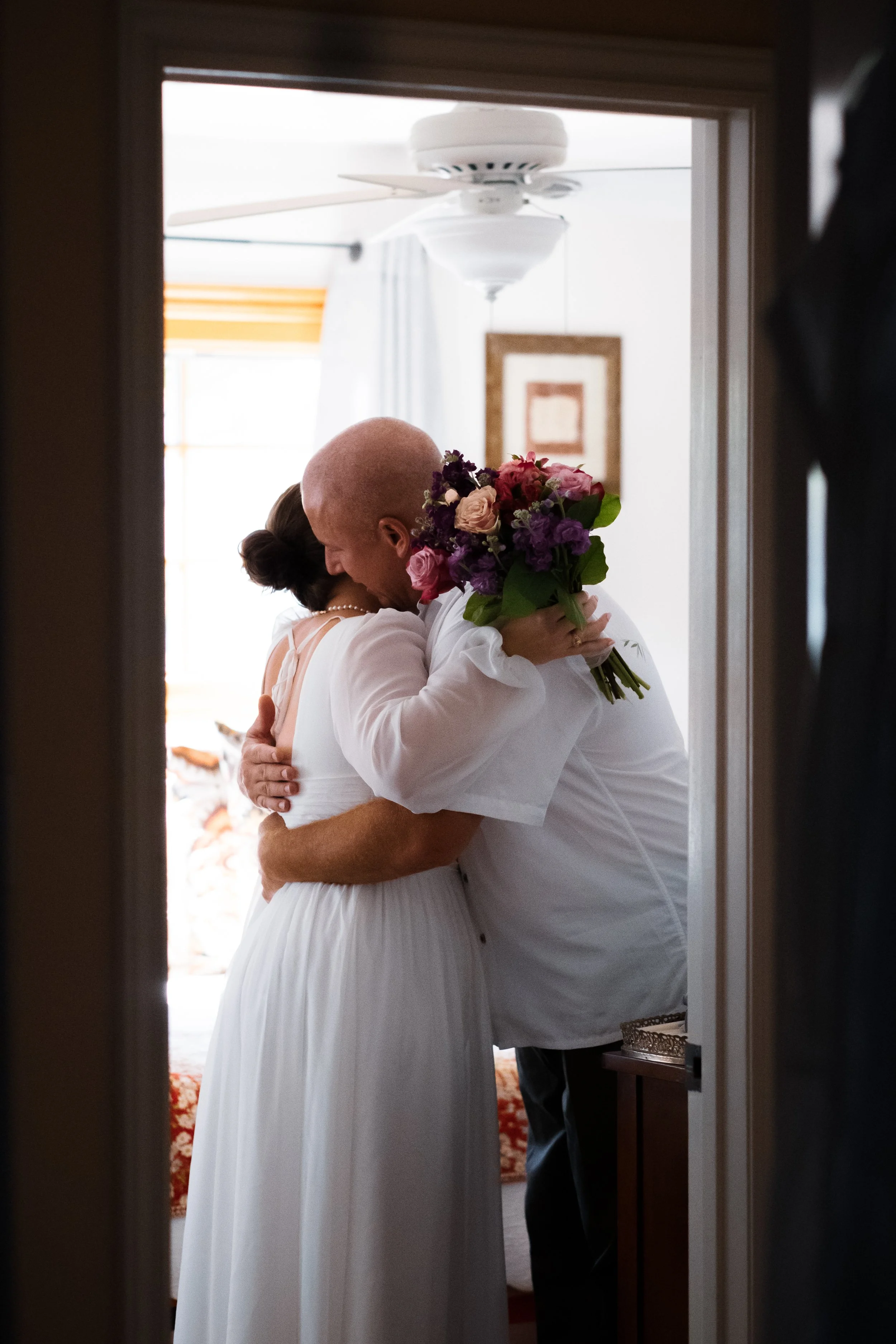 A couple embracing during their wedding in a room with a ceiling fan and framed artwork on the wall. The bride wears a white dress and the groom wears a white shirt. The groom holds a bouquet of pink and purple flowers.