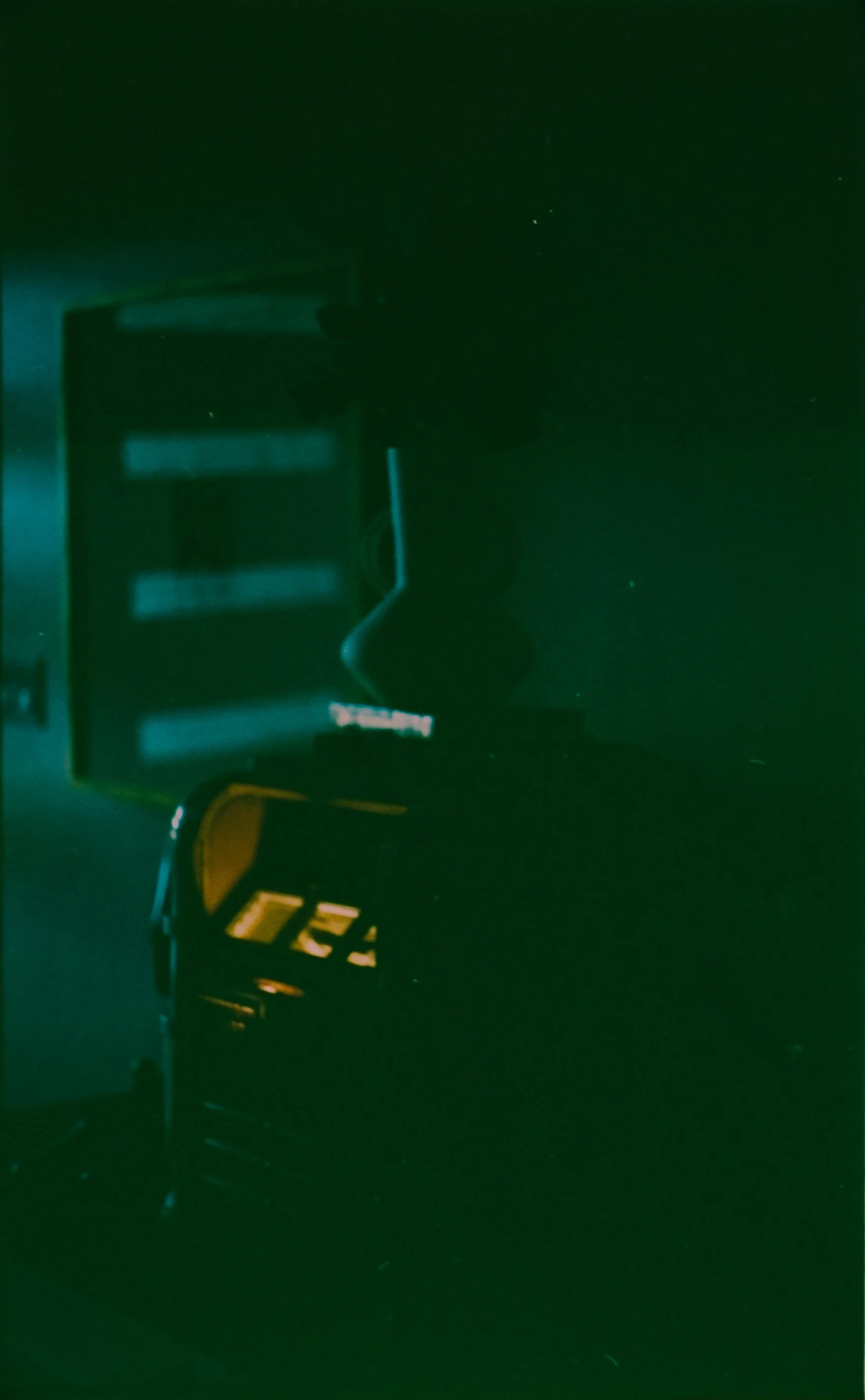 A dark, roughly square-shaped object with a greenish hue, reflecting a faint light source. The surface appears glossy and smooth, with some subtle reflections and slight imperfections visible.