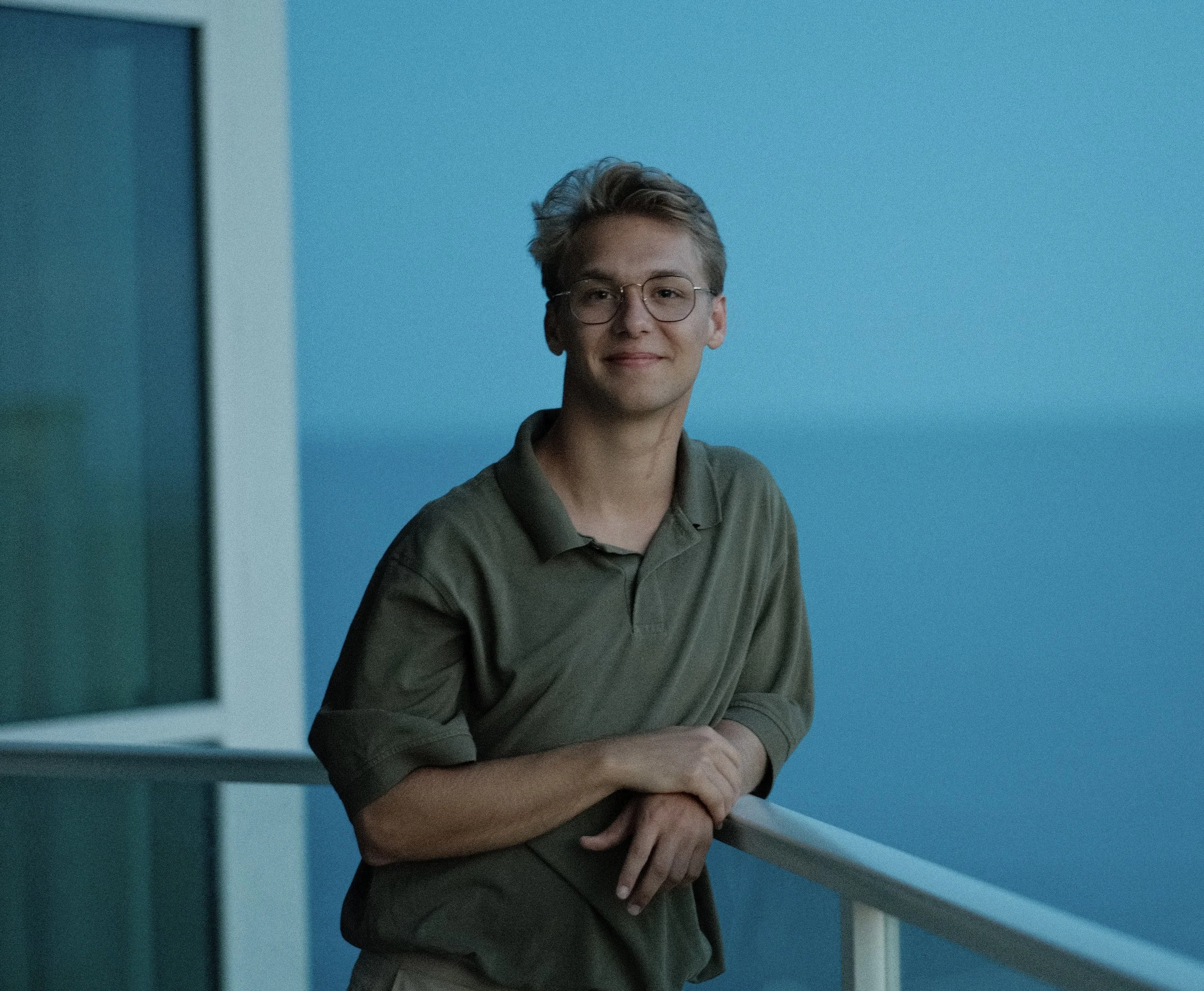 A young man with glasses and short, wavy hair stands with his arms crossed, smiling softly, on a balcony.