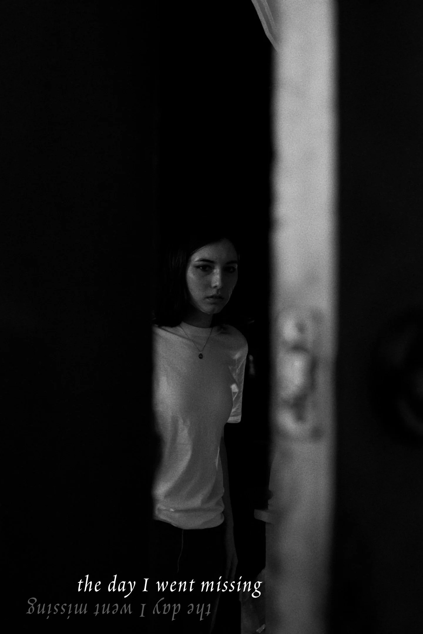 A young woman with dark hair and light skin wearing a light-colored t-shirt and a necklace with a small pendant, standing in front of a mirror behind a dark door or wall, with the text 'the day I went missing' reflected at the bottom of the image.