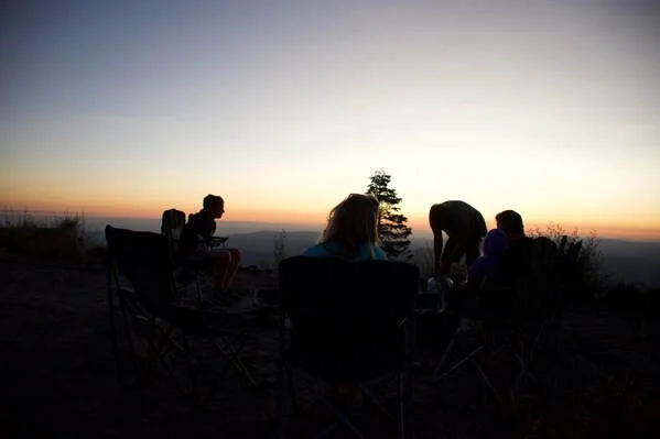 Group of people sitting around a fire pit on a hilltop at sunset with a view of the horizon.
