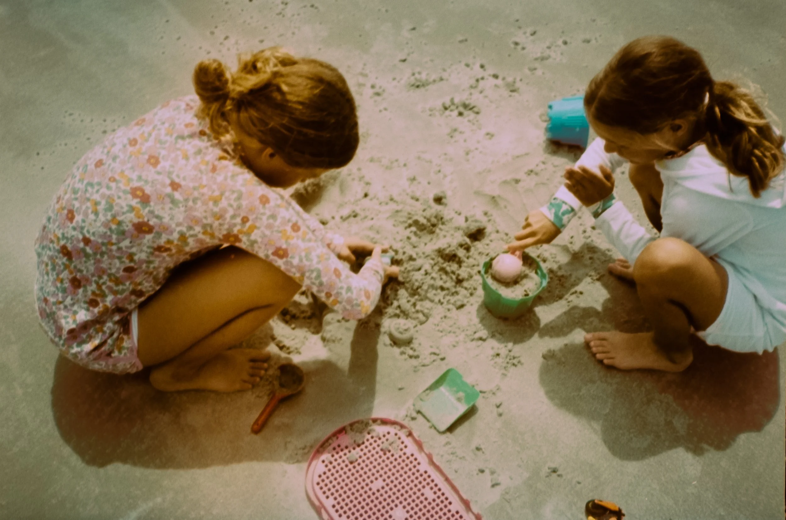 Two young girls playing in the sand indoors, surrounded by various toys and containers, with one girl wearing a floral dress and the other in a white outfit.