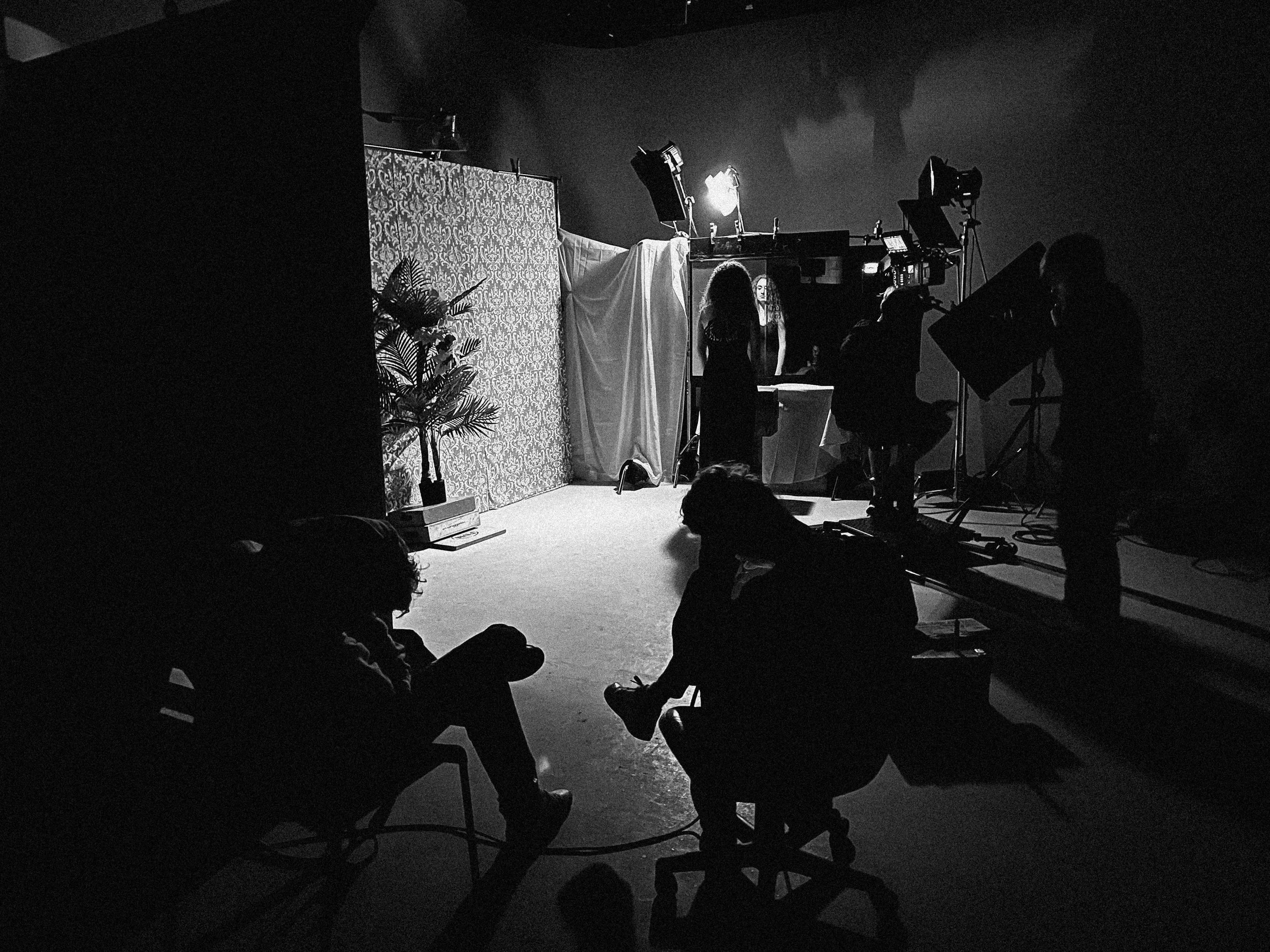 Black and white photo of a photo or film studio with people working on or near camera equipment, facing a backdrop. The silhouette of people are visible, and various lighting and filming equipment are set up.
