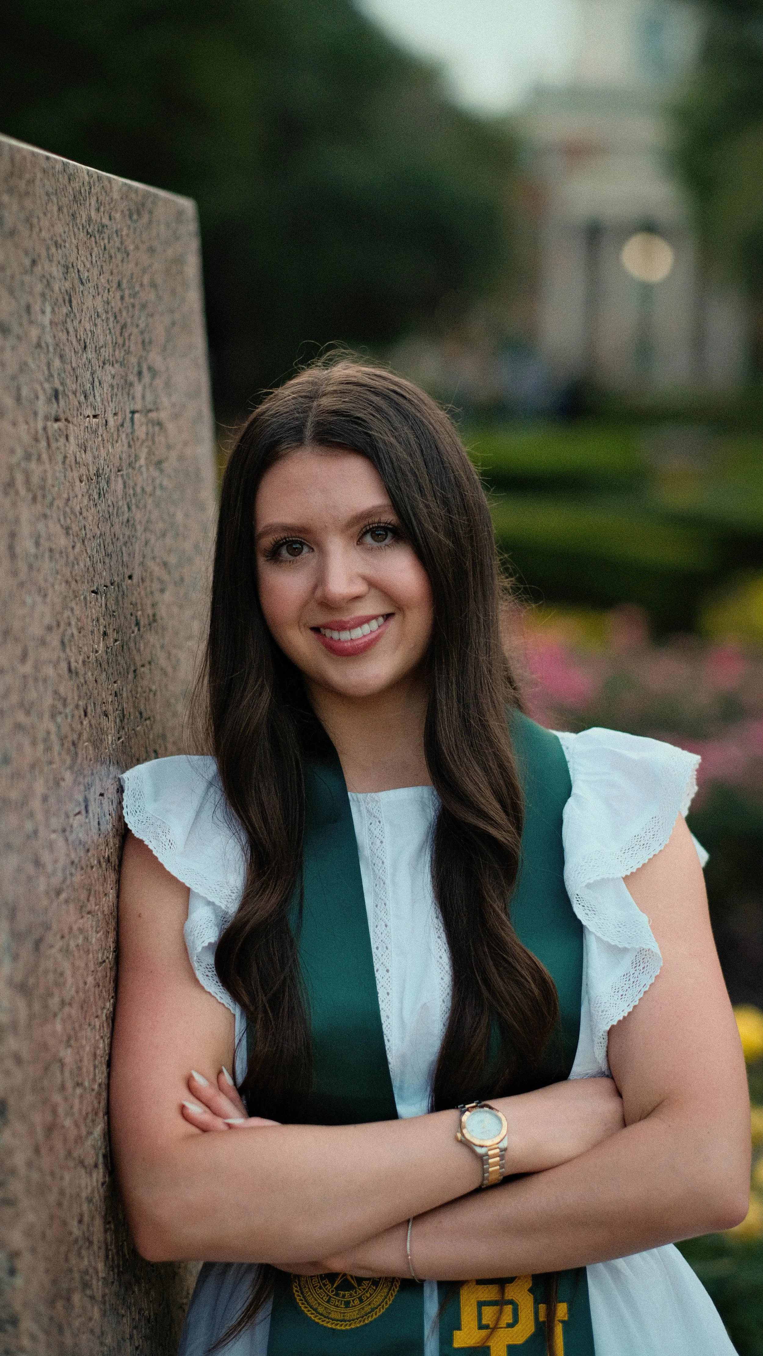 Young woman in graduation cap and gown smiling, leaning against a stone wall outdoors with blurred greenery in the background.