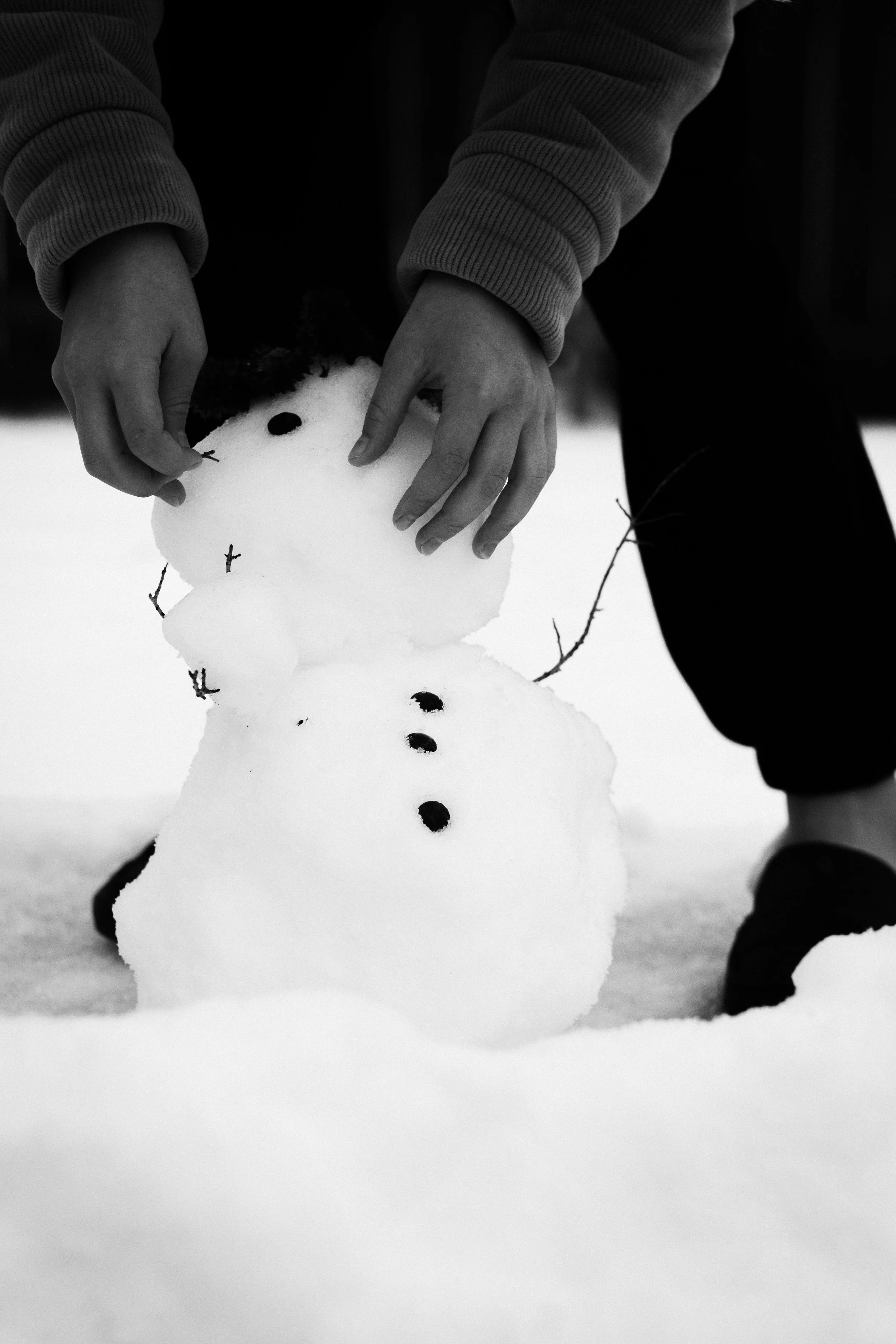A person building a snowman outdoors in a snowy landscape, with the snowman having a small face and a row of stones for buttons.