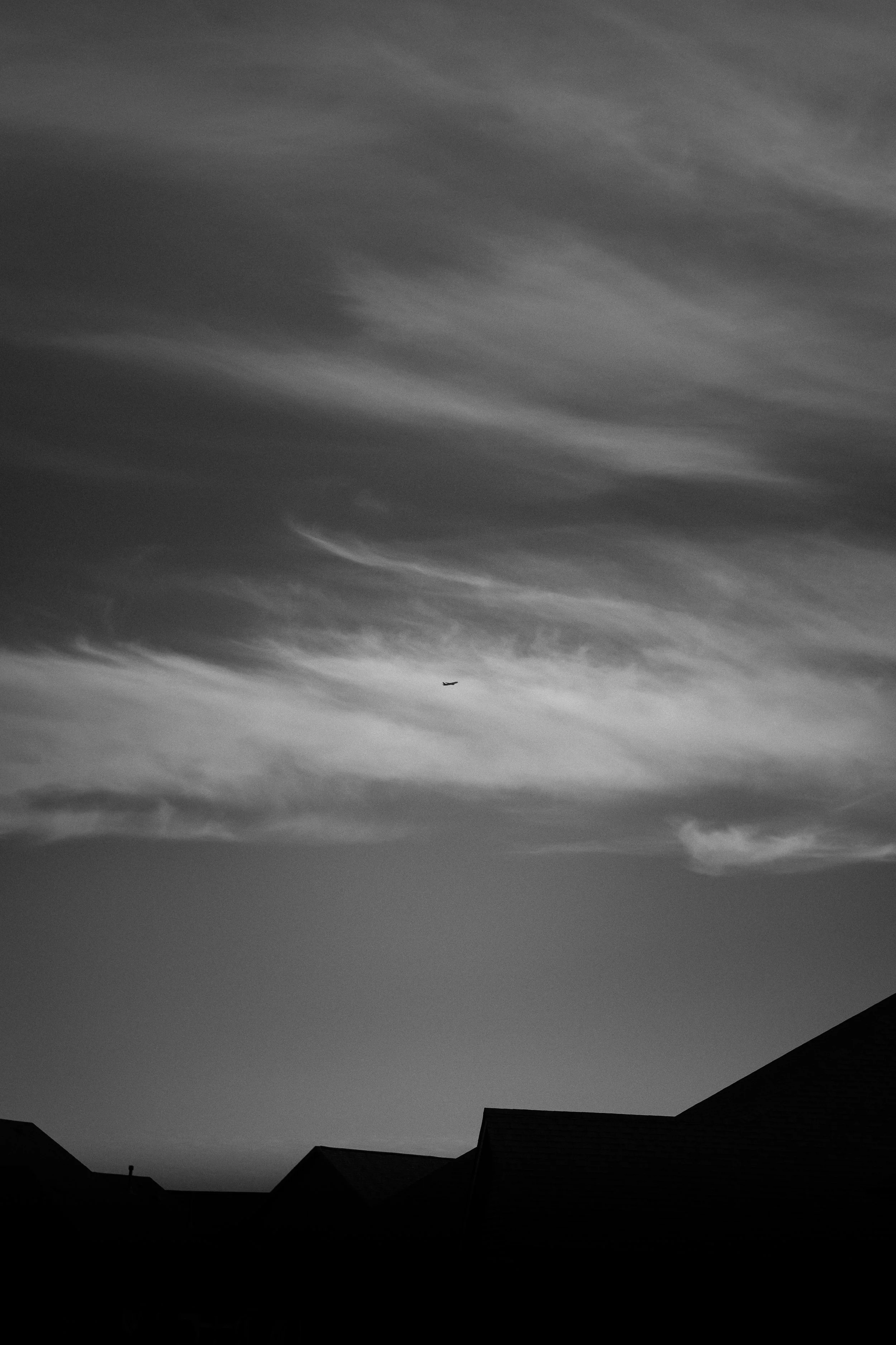 Black and white photo of a sky with wispy clouds and a small airplane flying in the distance above rooftops.