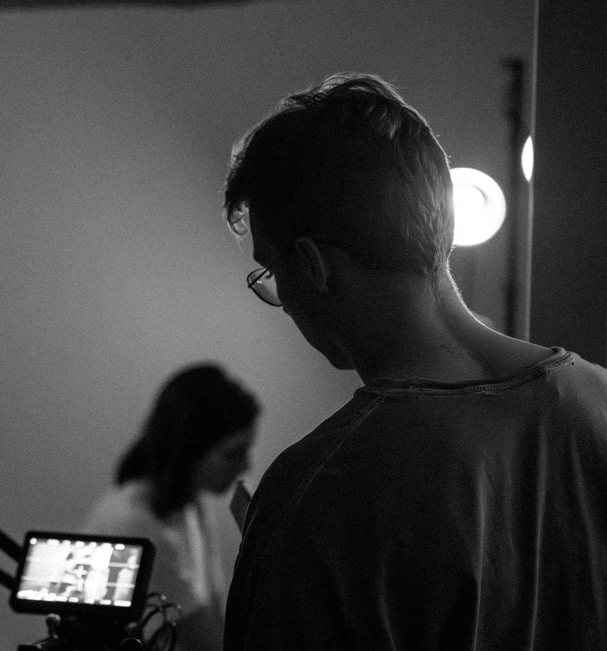 A black and white photo of a person with glasses and short hair, directing a performance in a film, with another person blurry in the background.