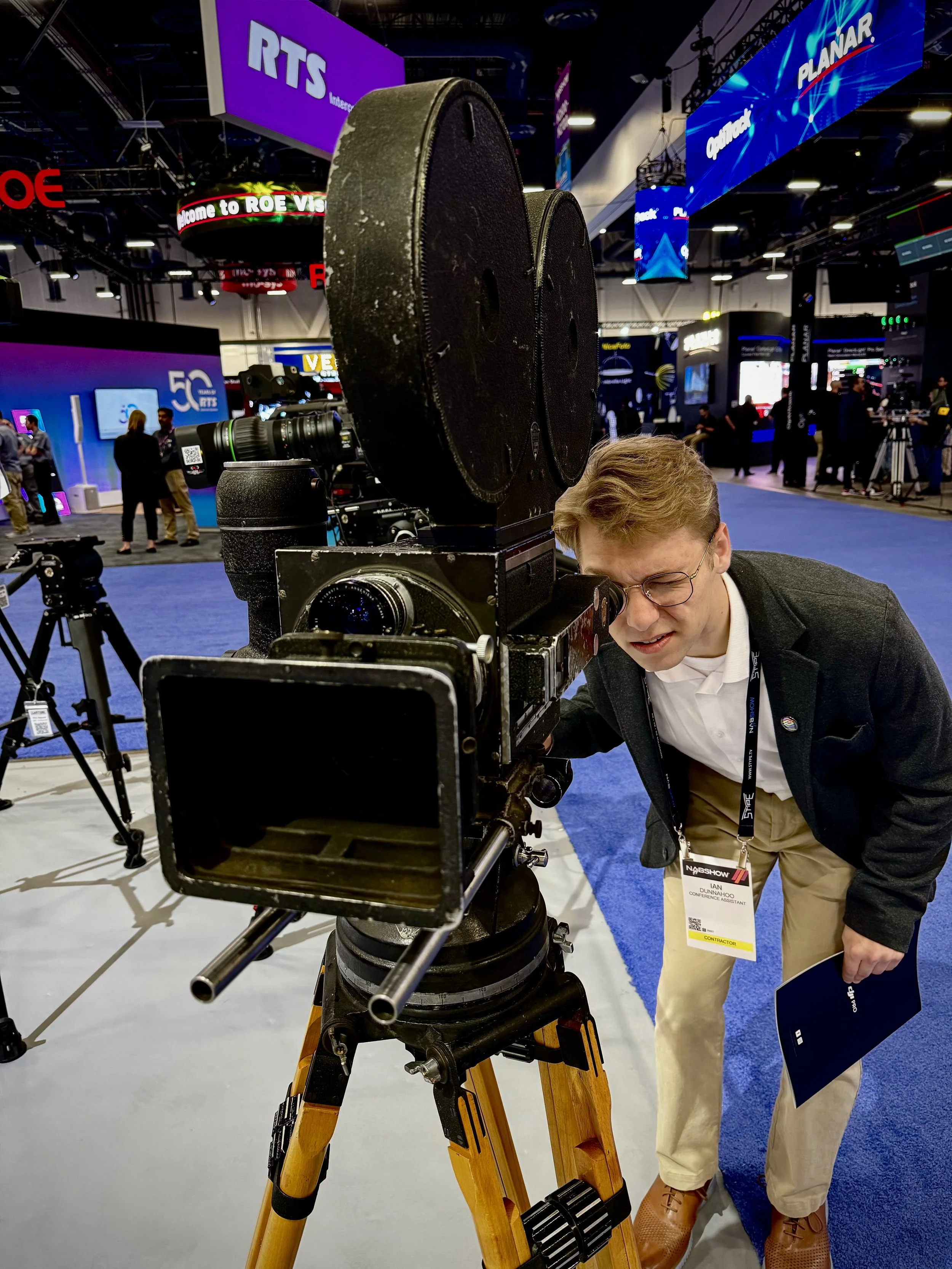 A man with glasses and a conference badge examining a large professional camera setup on a tripod at an indoor technology exhibition.