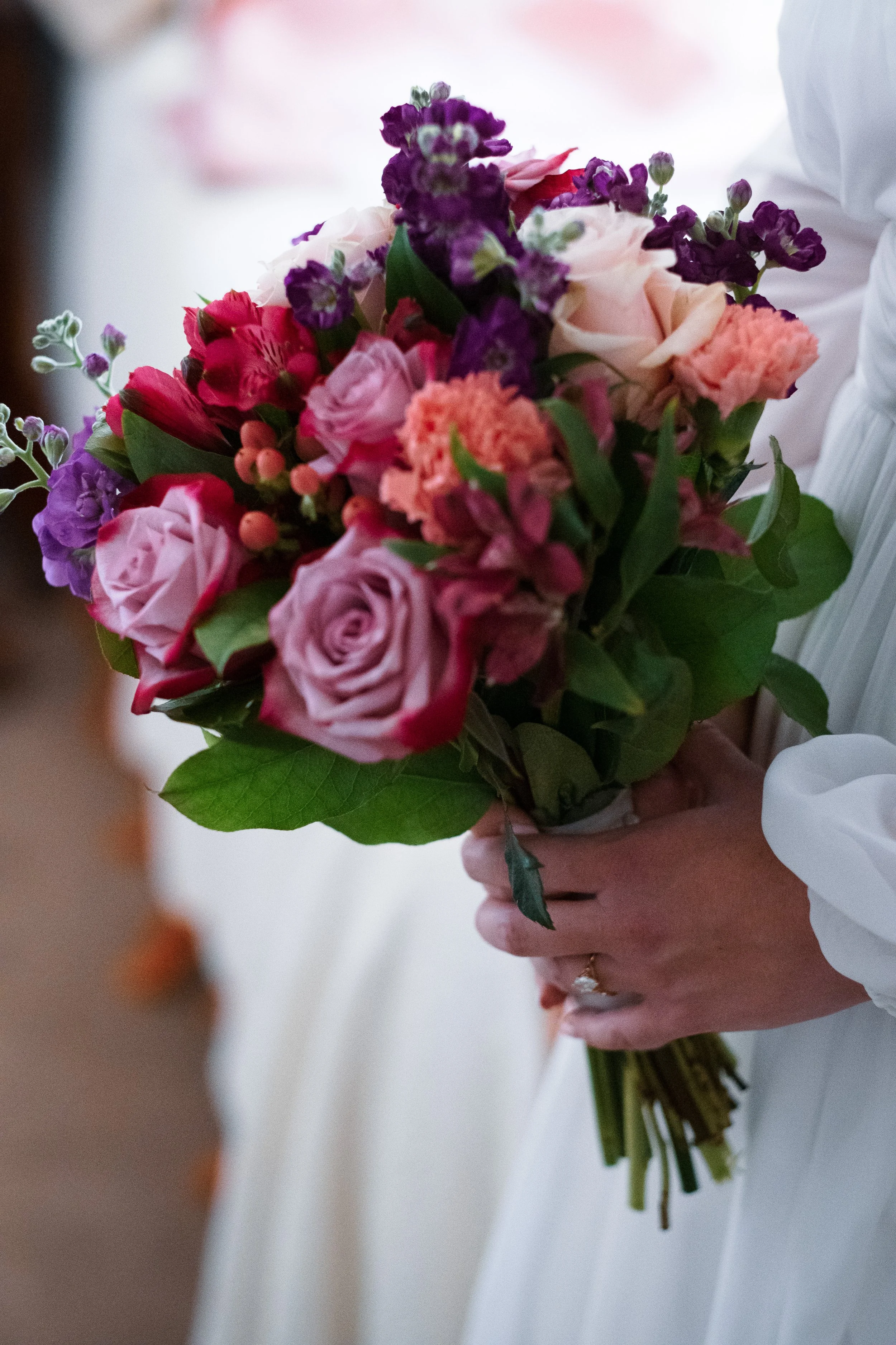 A person holding a colorful bouquet of flowers including pink roses, purple flowers, and green foliage.